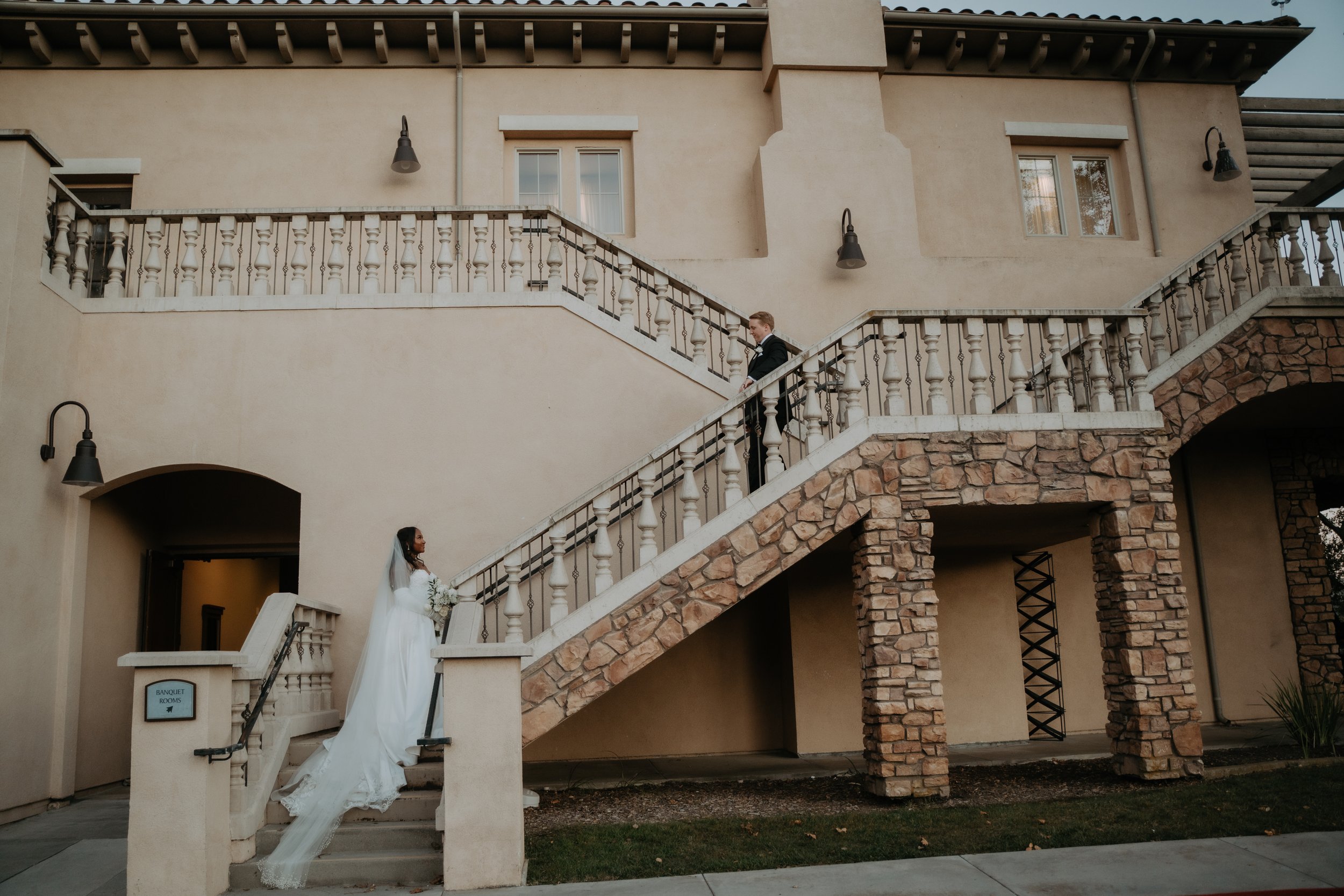 A bride in a white wedding dress holding a bouquet standing at the bottom of an outdoor staircase, looking up at a groom in a black suit standing on the top level of the staircase, with a stone and stucco building in the background.