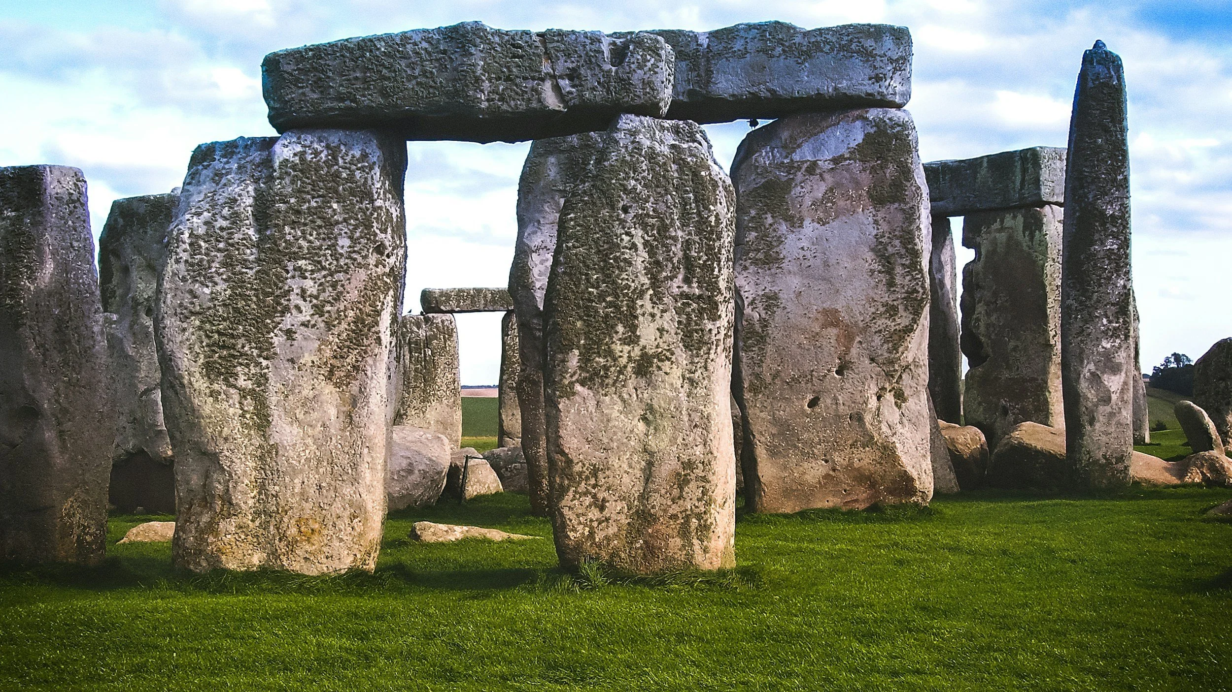 Stonehenge, a prehistoric stone circle with large upright stones topped with horizontal lintels, set in green grass under a cloudy sky.