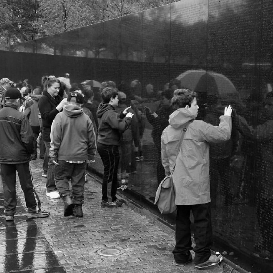  students are standing at the Vietnam Veterans Memorial and touching wall to feel engraved names in the rain in black and white photo  Students learn about the men and women that died or were missing in action during the Vietnam War and remember them