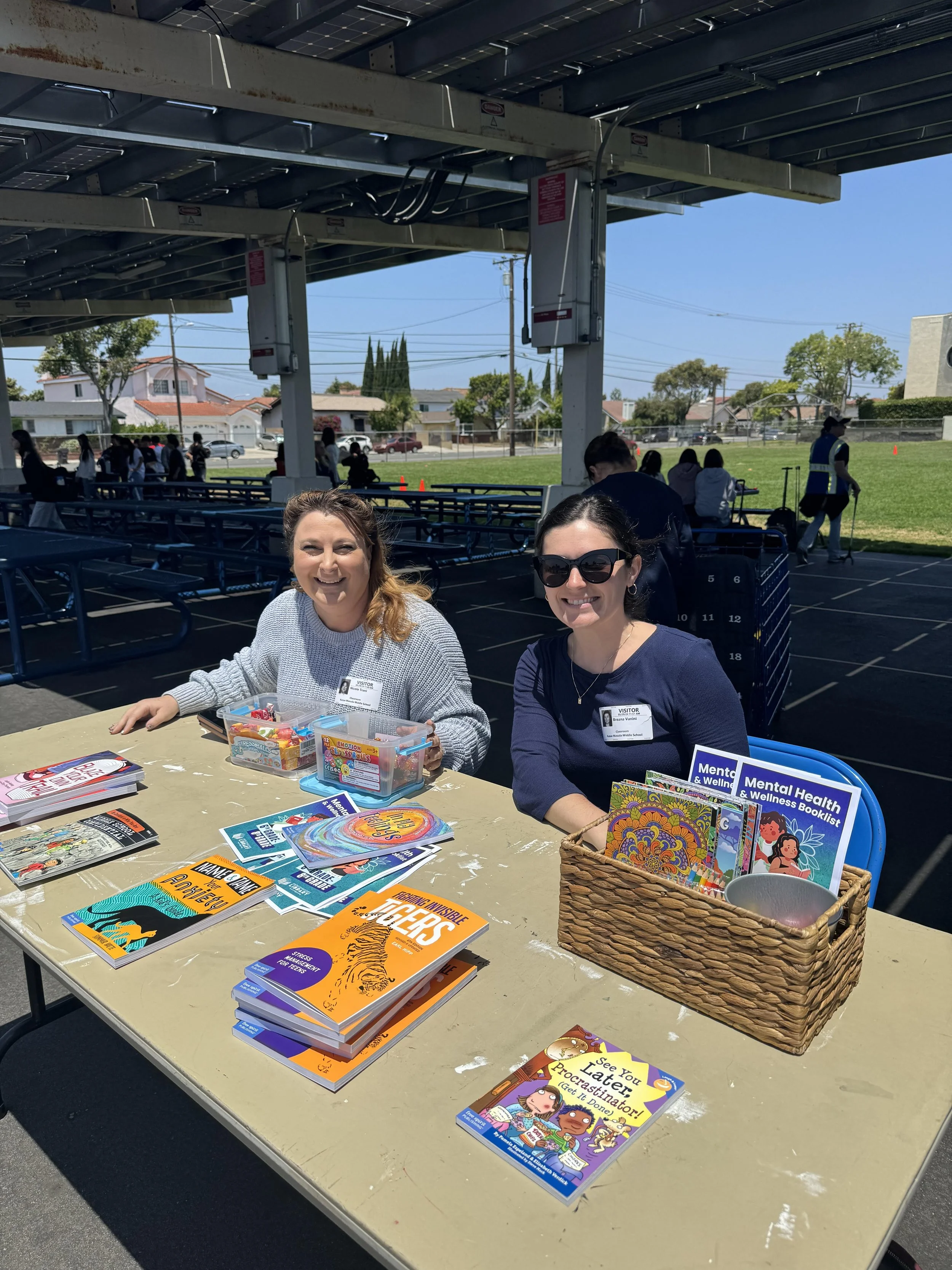 Two women are sitting at a table outdoors with various mental health and wellness books, pamphlets, and promotional materials displayed. One woman is smiling and holding a container of candy, while the other woman, wearing sunglasses, is smiling at the camera. In the background, there is a grassy area with people and structures under a partly cloudy sky.