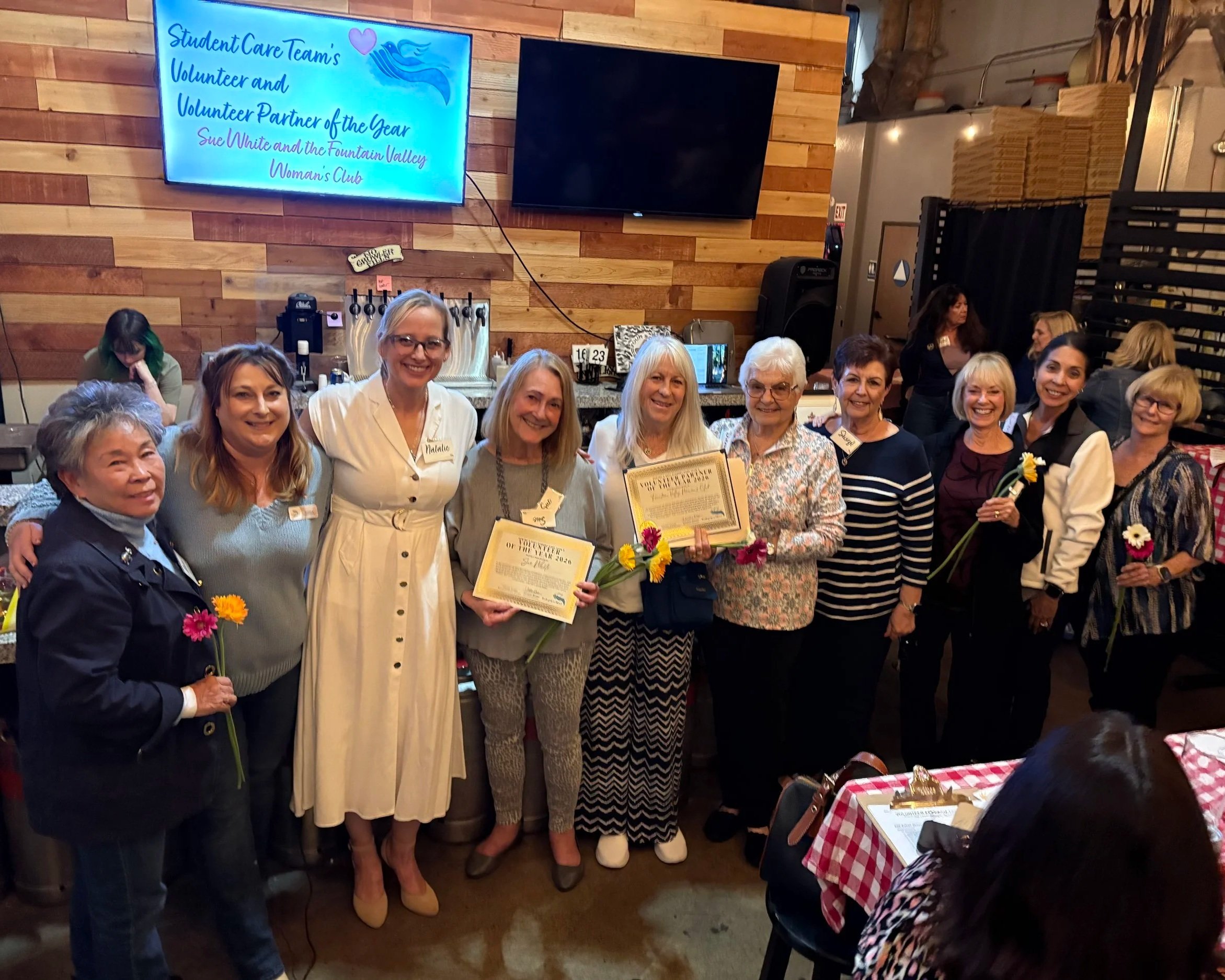 A group of women gathers together, some holding flowers and certificates, at an indoor event with a wooden wall in the background. A large screen displays a message about an award for Volunteer of the Year, and some women are dressed in semi-formal attire. The setting appears to be a celebration or recognition event.