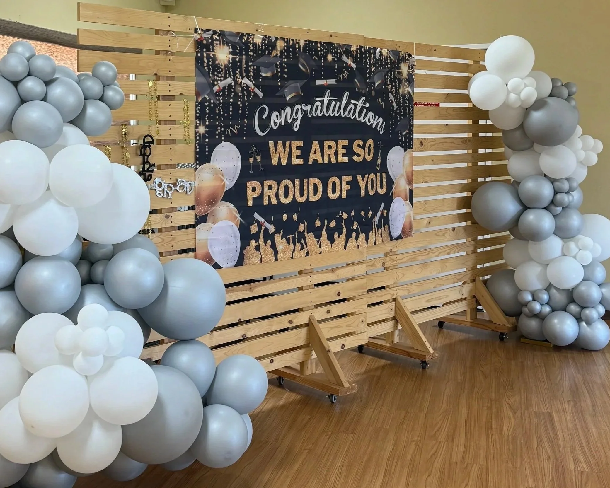 Decorative backdrop with the words "Congratulations, we are so proud of you" on a black banner, surrounded by white and grey balloons, with a wooden slat background.