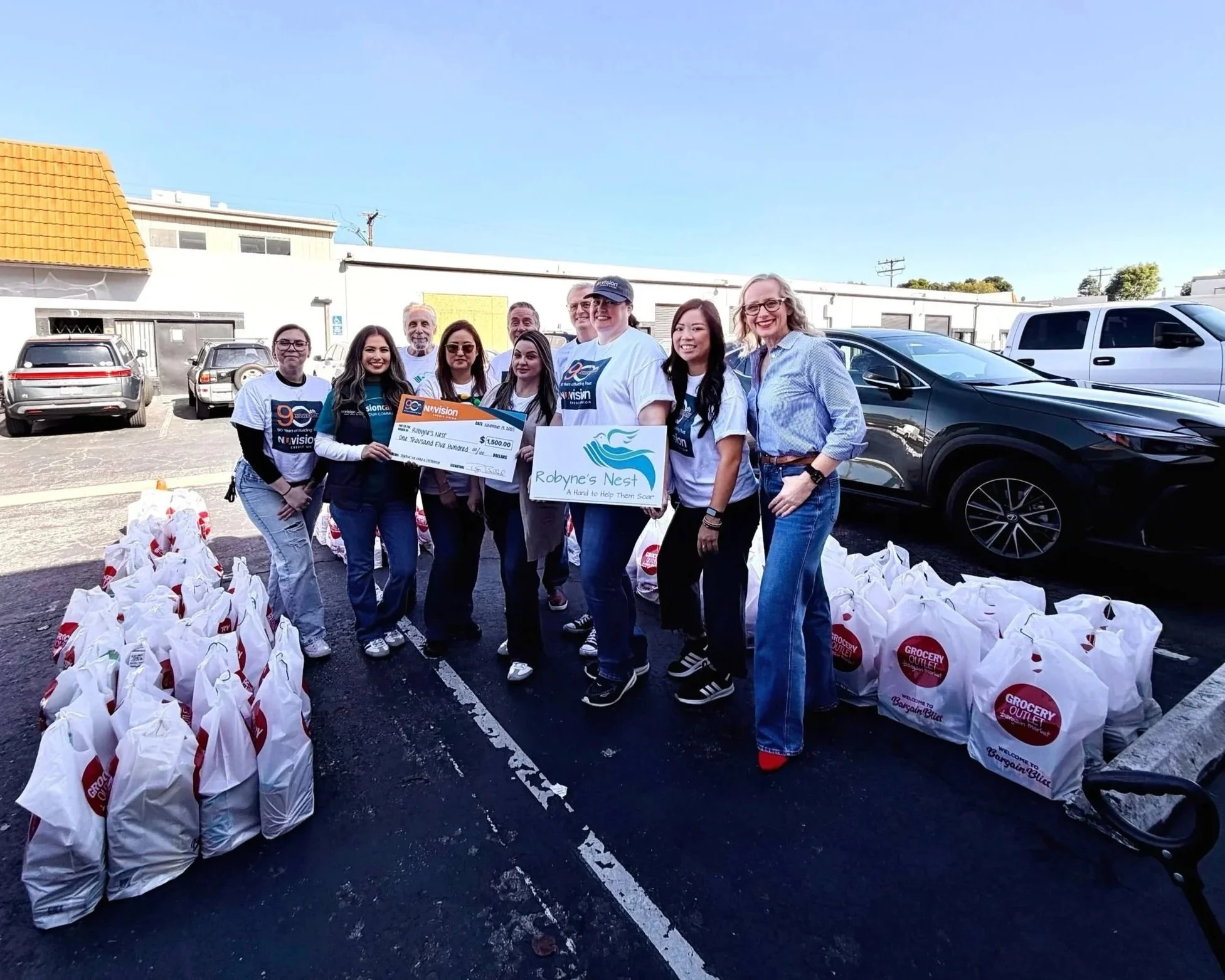 Group of people standing in a parking lot, smiling, with grocery bags and a large check for $1,500, and a sign for Robyne's Nest, a charity organization, in front. The background includes parked cars and a building.