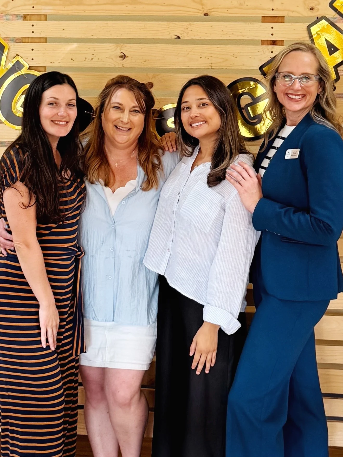 Four women standing together, smiling and posing for a photo, in front of a wooden wall decorated with gold and black balloons.