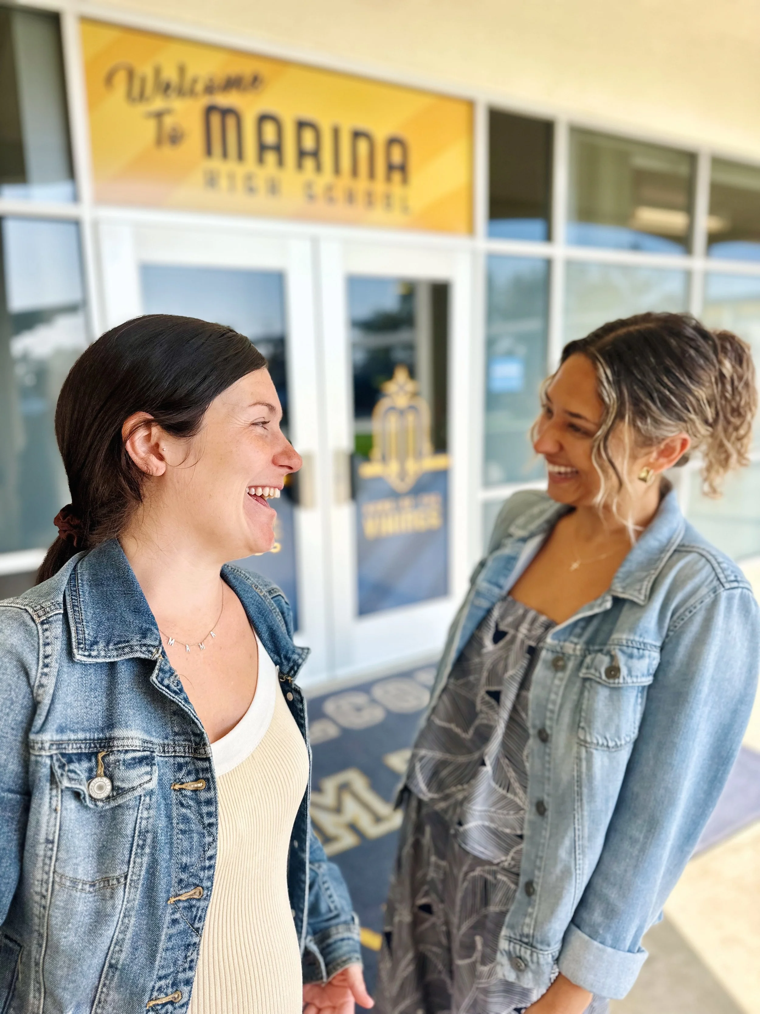 Two women smiling and facing each other in front of a yellow sign that says 'Welcome to Marina Regional School.'