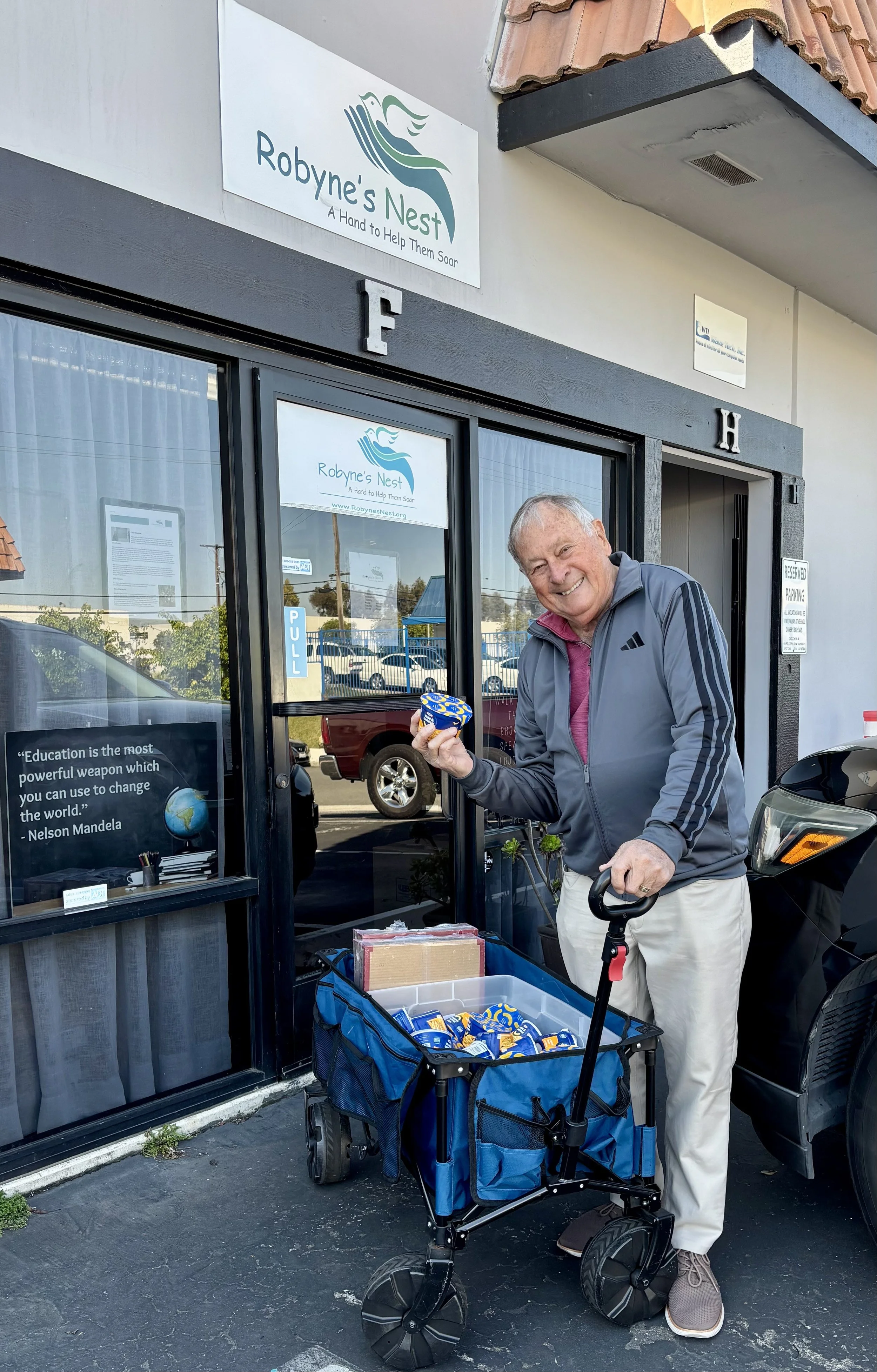 An elderly man outside of a building with a sign that reads "Robyne's Nest." He is smiling and holding a container of ice cream, with a walker nearby loaded with supplies. There is a quote on the window about education by Nelson Mandela.