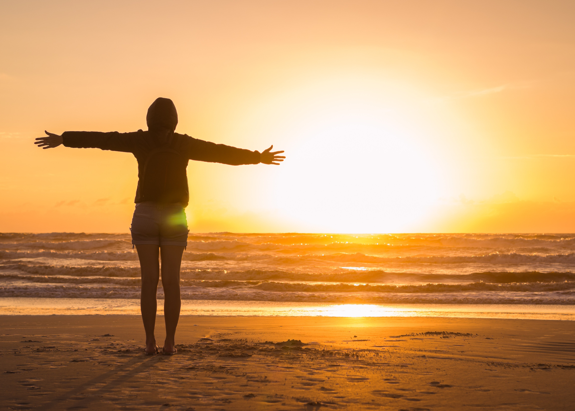 Person with arms outstretched on a beach at sunset.