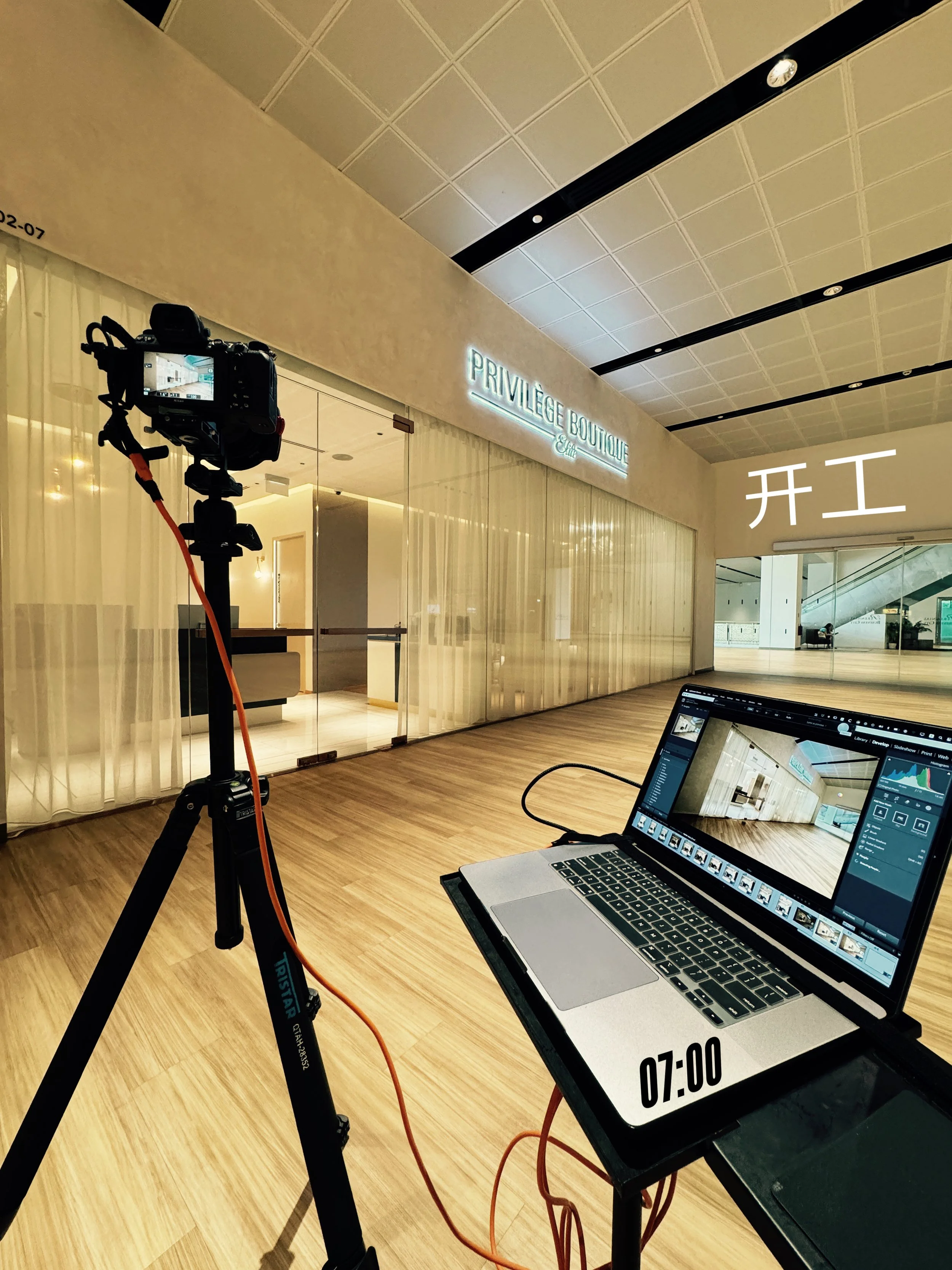 Photo of an indoor space with a sign reading 'PRIVILEGE BOUTIQUE.' A tripod with a camera is set up, and a laptop is displaying the same space with a timestamp '07:00.' There are curtains covering glass walls and an escalator visible in the backgroun