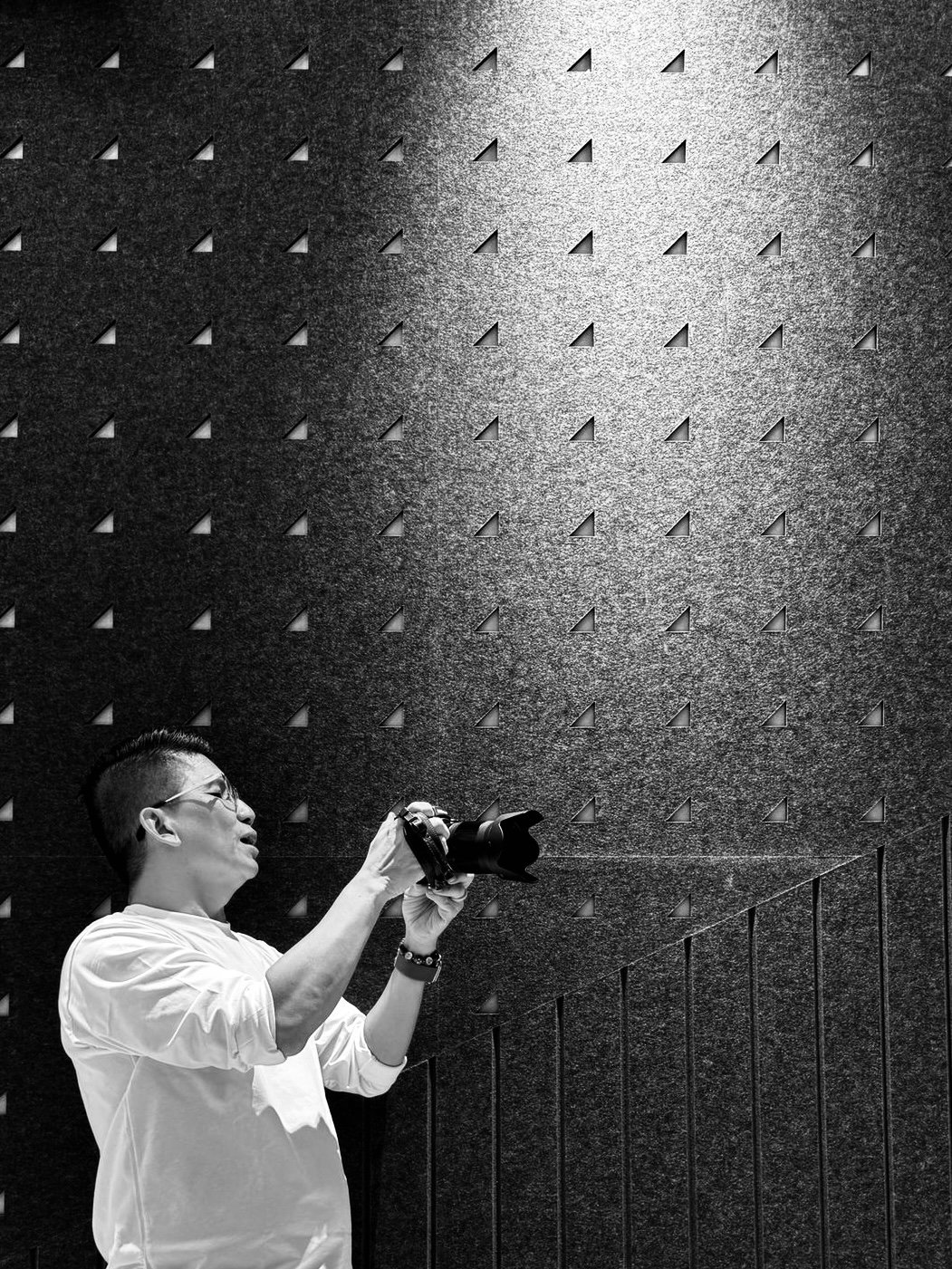 A man with glasses wearing a white long-sleeve shirt holding a camera, standing in front of a textured wall with small triangular cutouts and a diagonal line pattern.