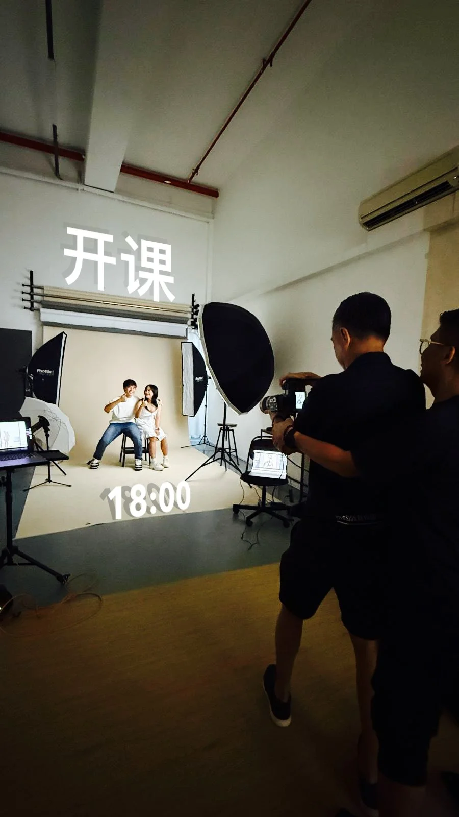 Studio photography setup with two people sitting on chairs in front of a beige backdrop, surrounded by large photographic lights, with two crew members operating a camera, and Chinese characters '开课' and a time '18:00' overlaid on the image.