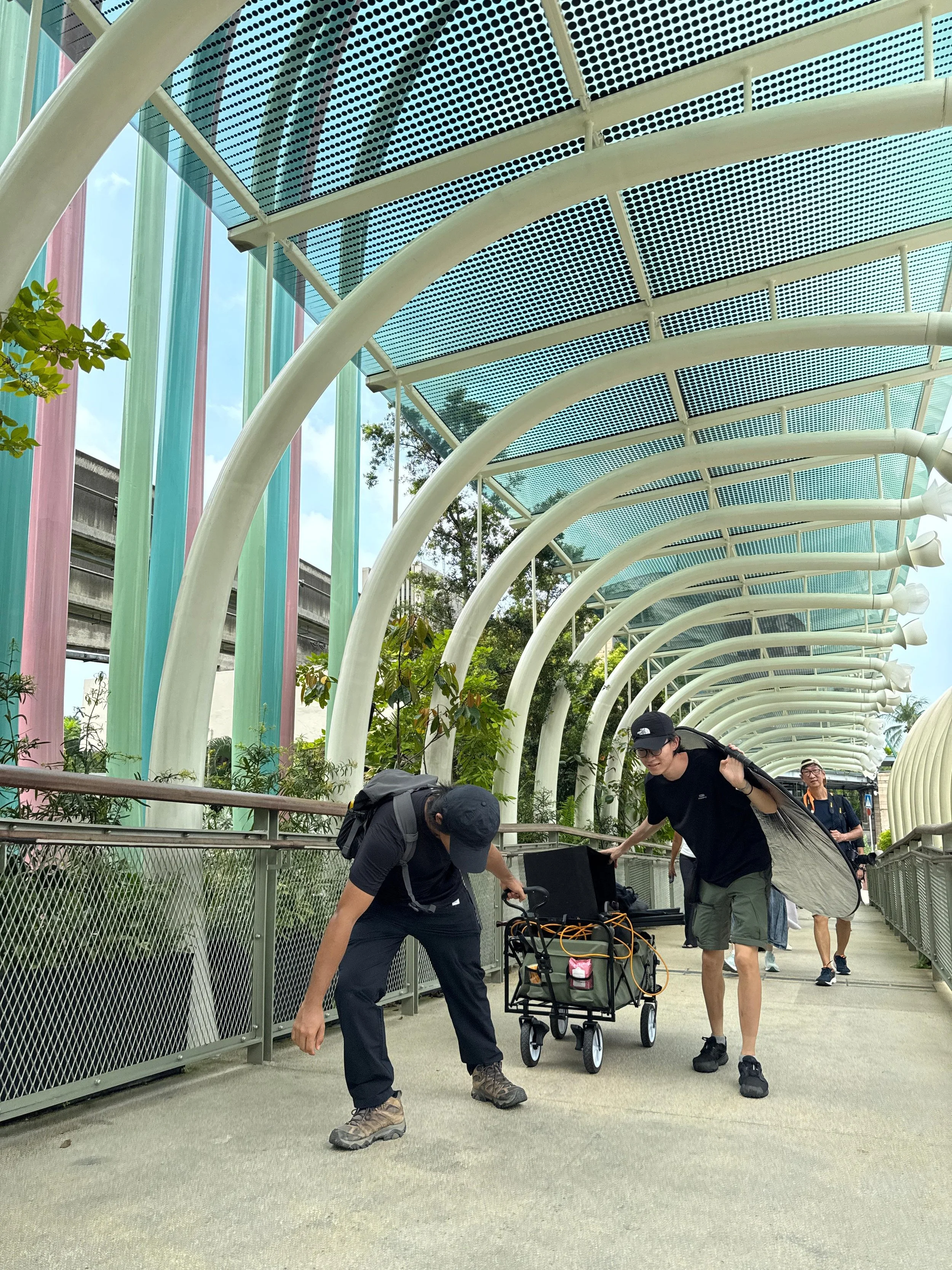 Two people adjusting equipment on a rolling cart on a covered walkway with colorful vertical pillars and curved white beams overhead.