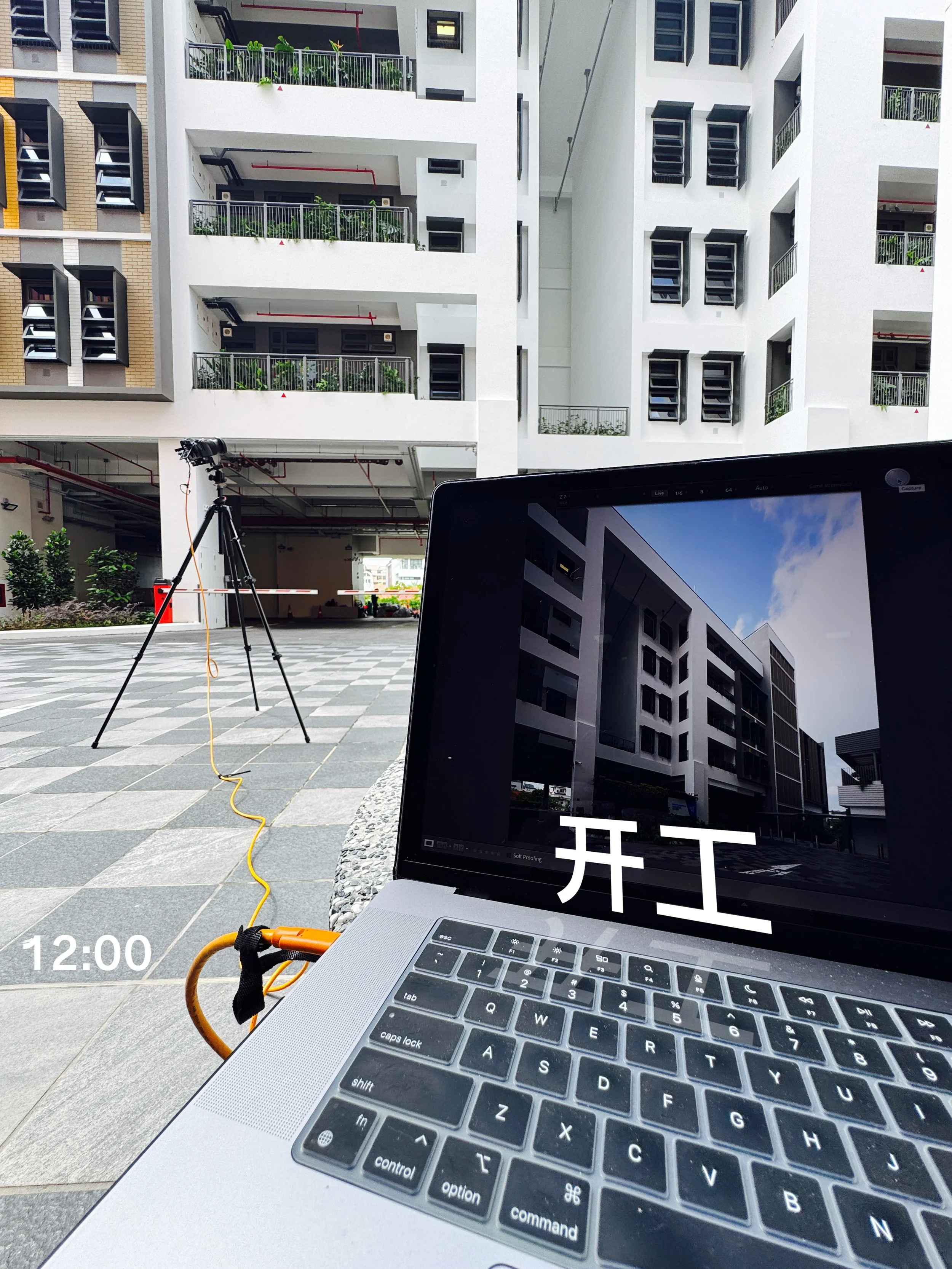 A laptop with Chinese characters on the screen, displaying a modern building, and a camera mounted on a tripod outside in a courtyard area, with other buildings in the background.