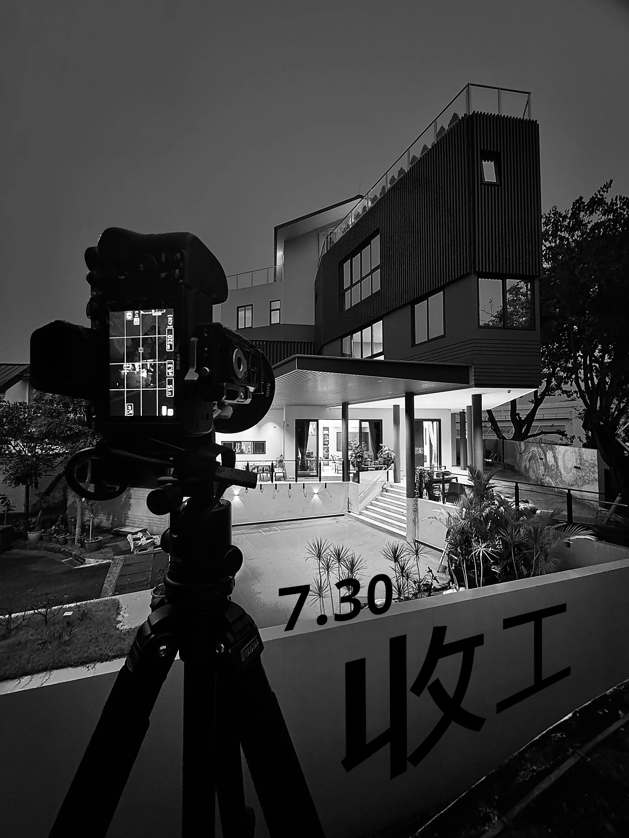 Black and white photo of a modern building during dusk, with a camera on a tripod in the foreground and a sign with the time 7:30 and Japanese characters.