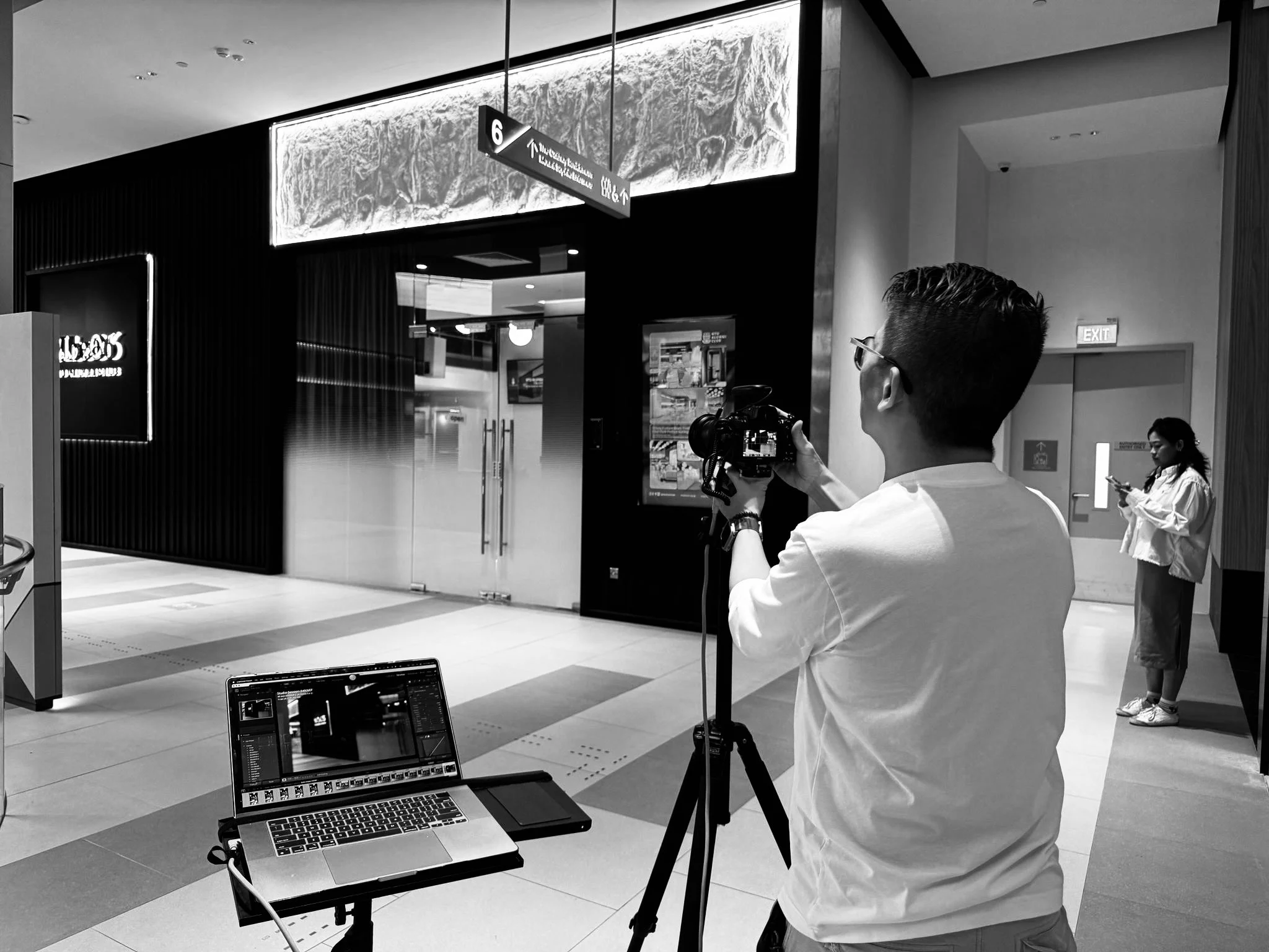 A person taking a photo with a camera mounted on a tripod in an indoor setting, with a laptop on a table nearby, and a woman looking at her phone in the background.