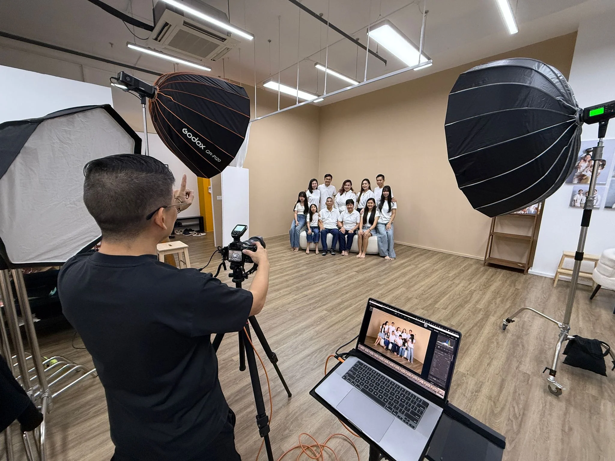 Photographer in a studio taking a group photo of 13 people, all dressed casually, with professional photography equipment including large softbox lights and a laptop displaying the photo editing software.