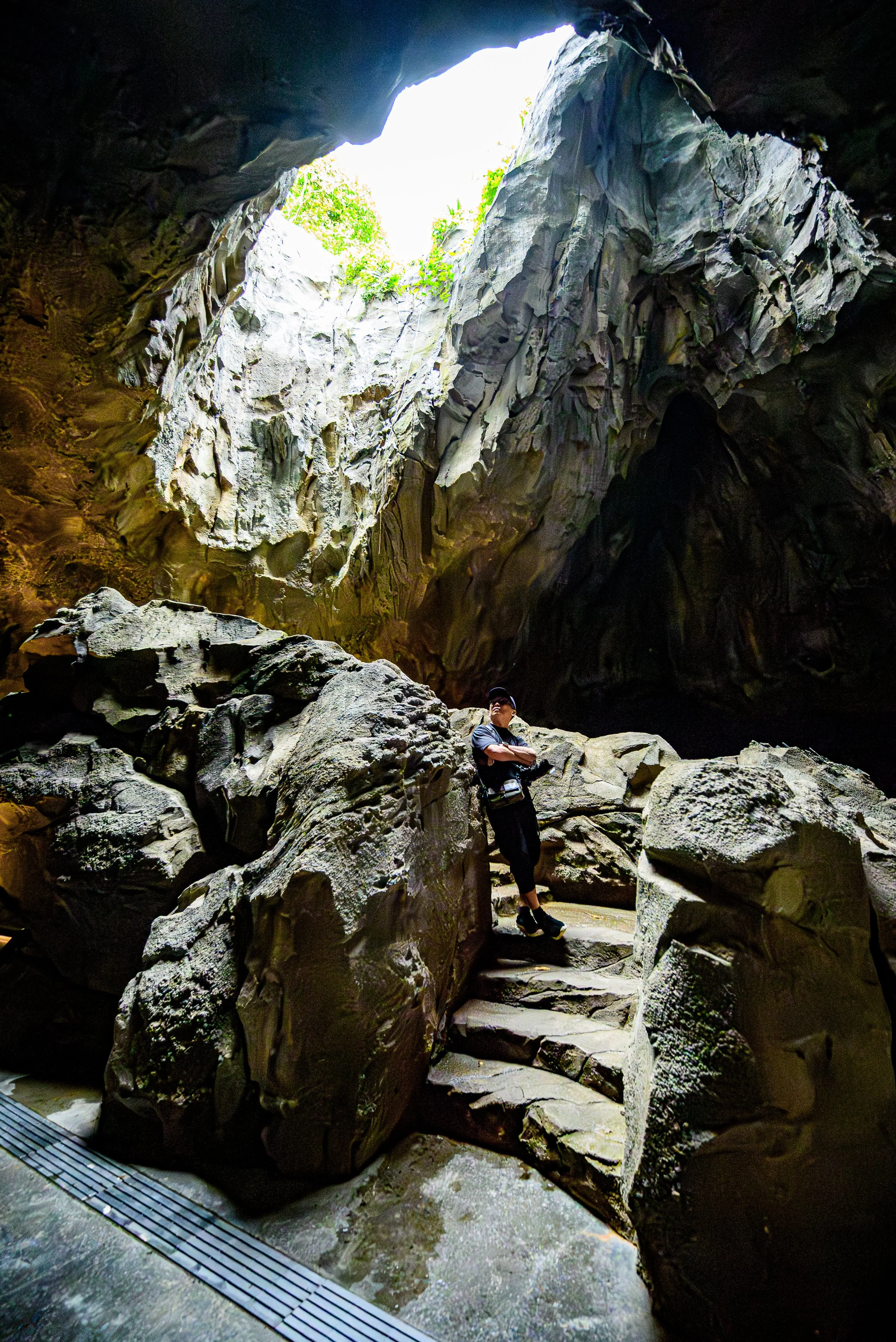 A person standing on a rocky ledge inside a large cave with a bright opening at the top, showing green foliage outside.