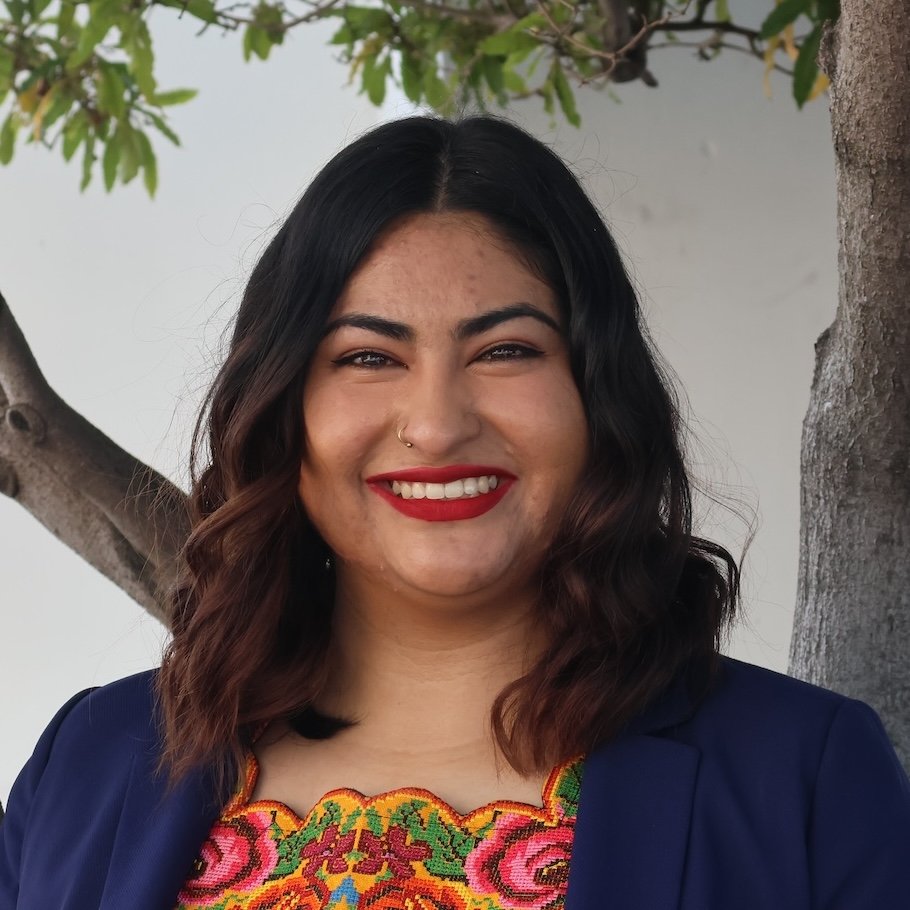 Headshot of Jessica Peregrina, who has wavy black and brown hair, wearing a floral top and a navy blazer