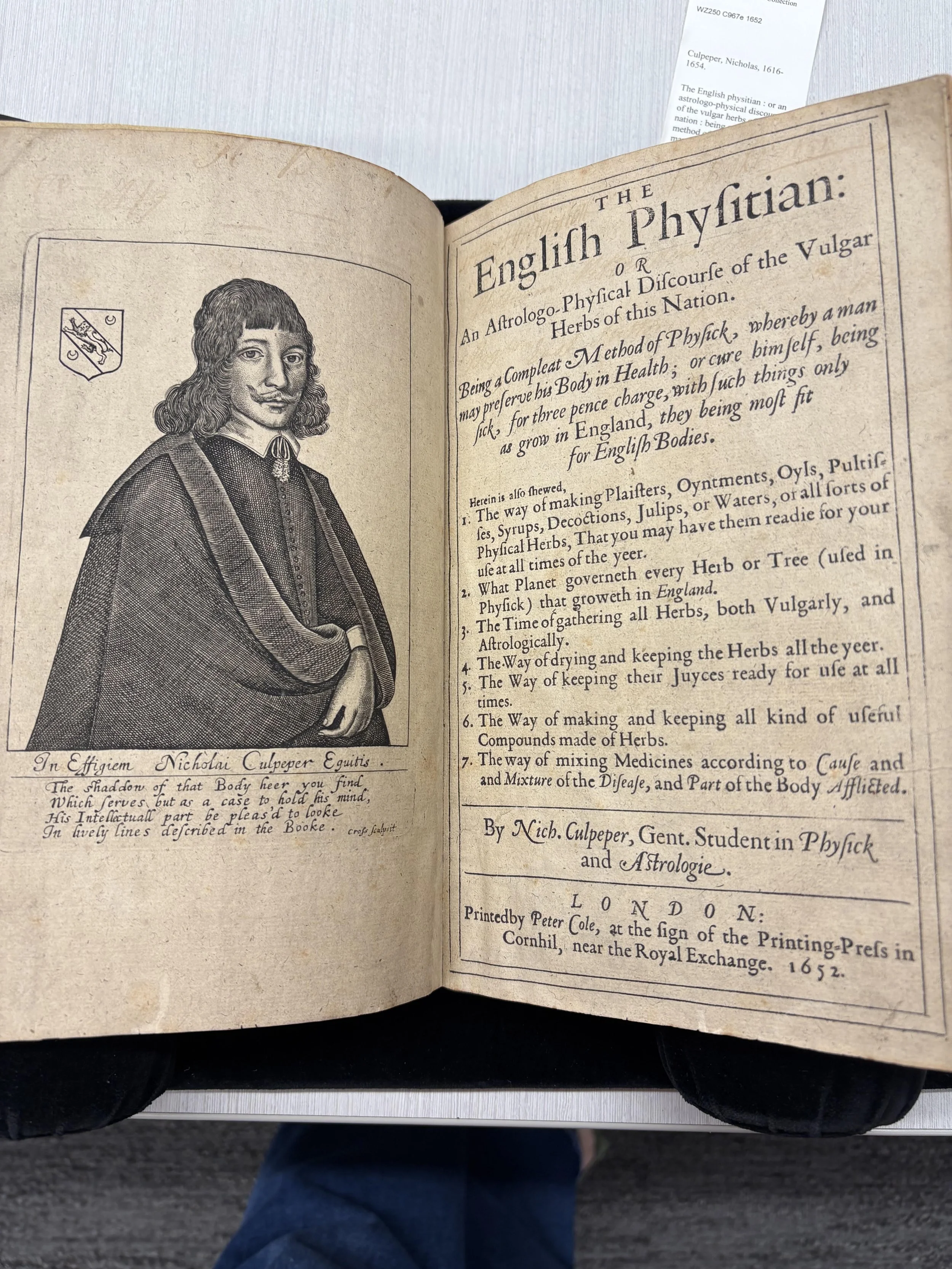 Open book showing a black and white portrait of a man with long, curly hair and a mustache, dressed in historic clothing, and a page with text titled "The English Physician" from 1652.