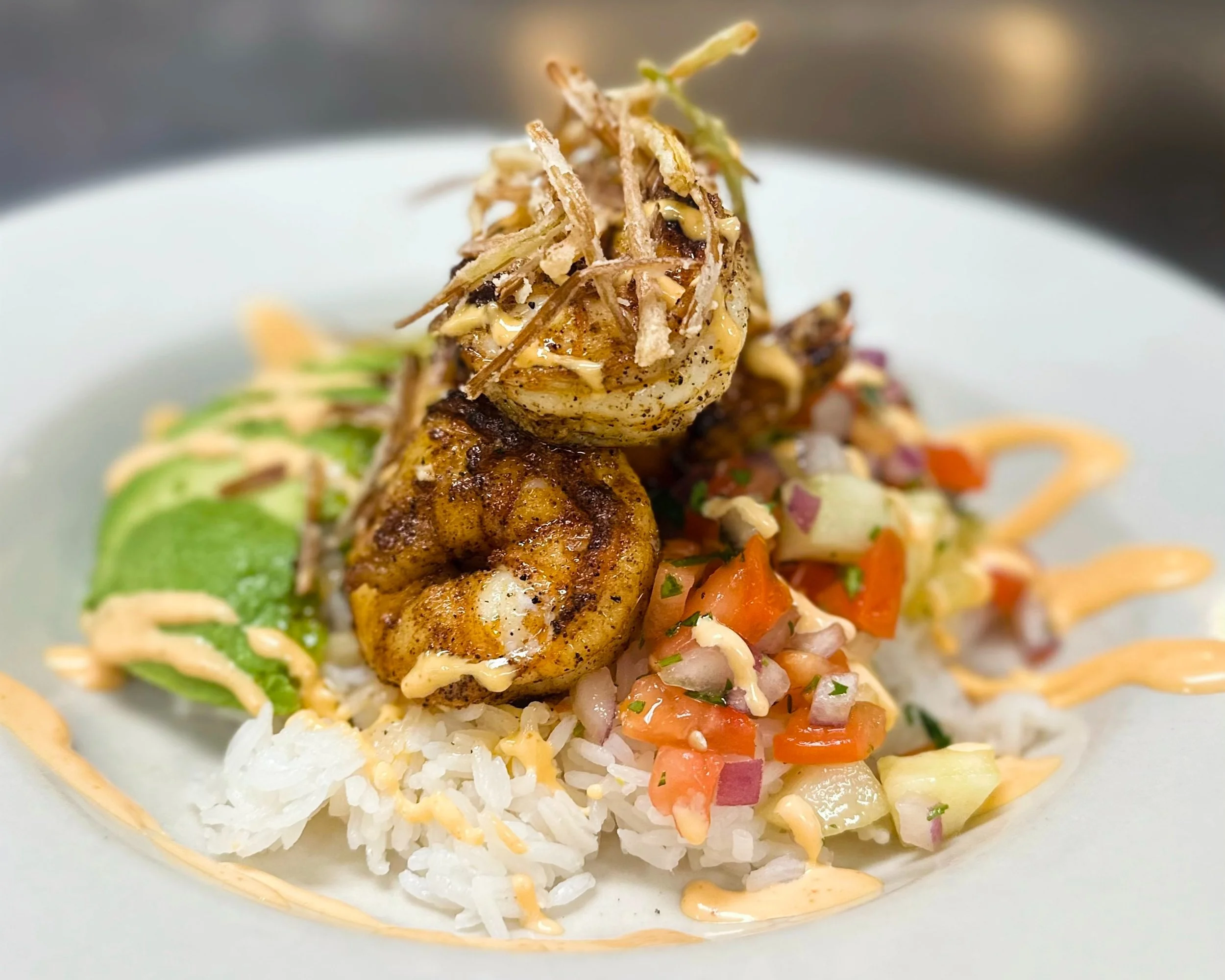 A plated dish featuring grilled shrimp atop rice, garnished with diced tomatoes, onions, avocado slices, and drizzled with an orange-colored sauce.