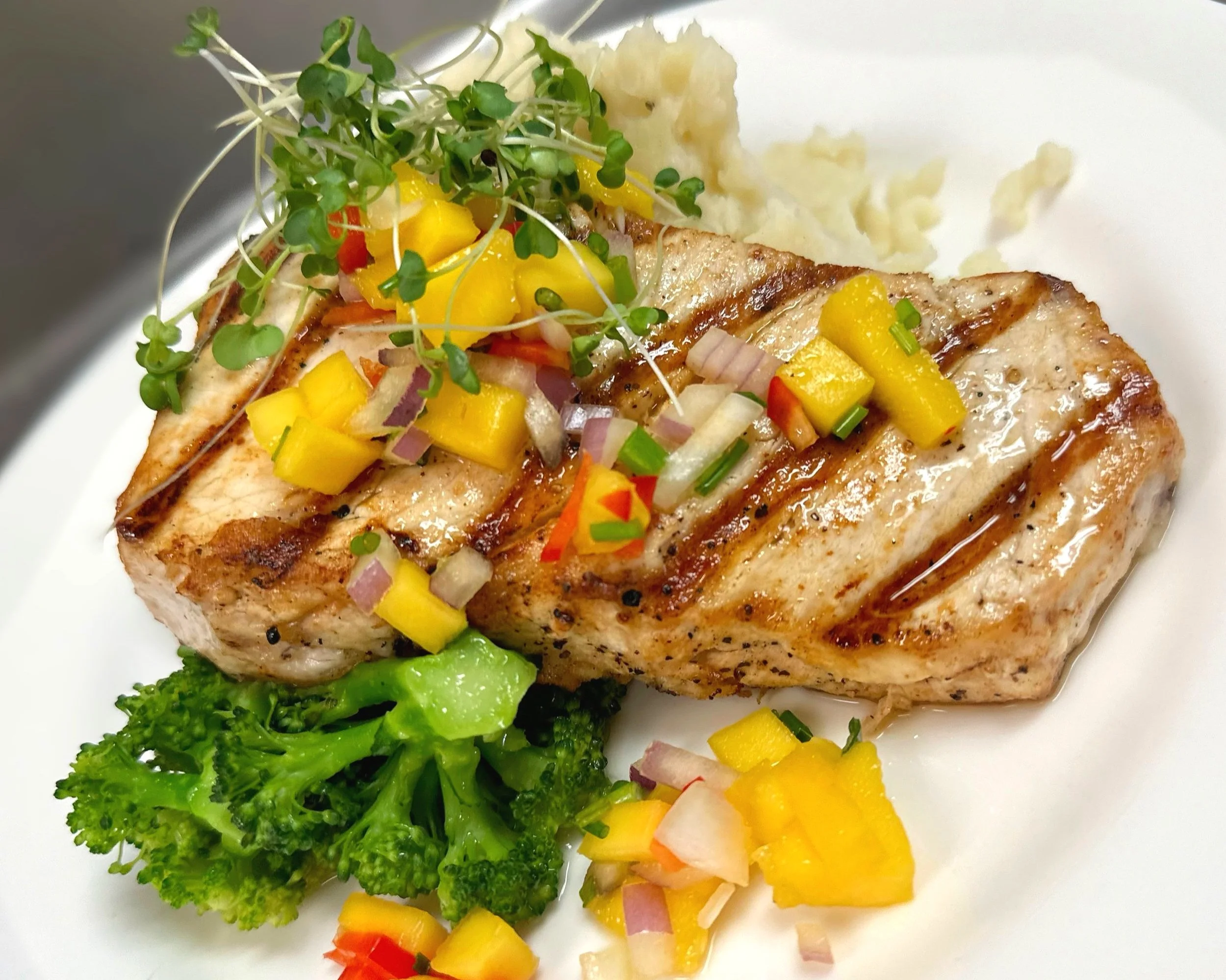 Grilled salmon fillet garnished with mango salsa, microgreens, and chopped onions, served with steamed broccoli and mashed potatoes.