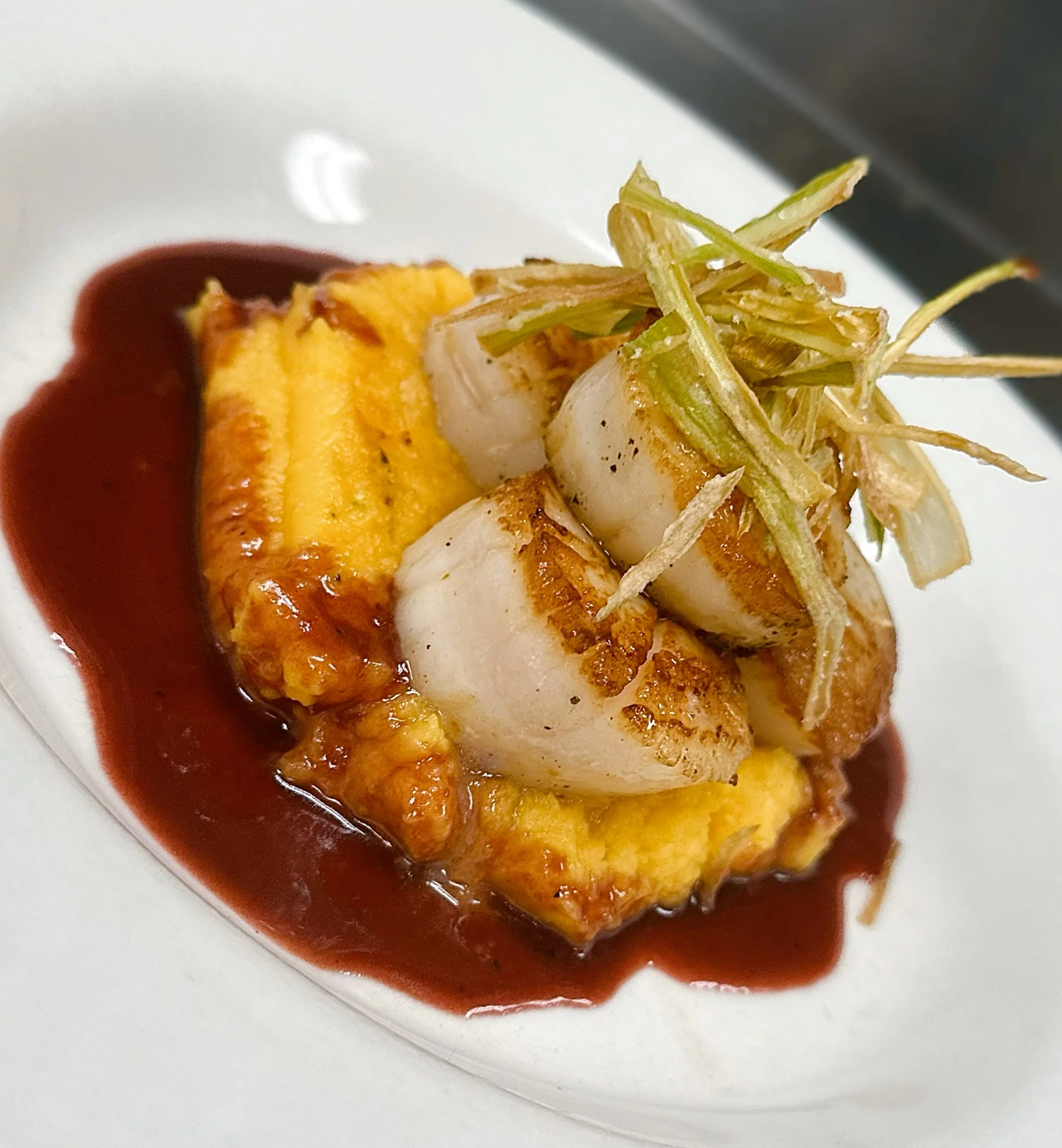 Seared scallops served on a bed of smooth mashed sweet potato with a red sauce, topped with shredded green garnish.