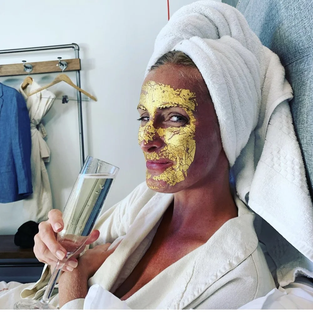 Carly Wahl in a white robe and towel turban, wearing a gold leaf face mask
              and holding a glass of champagne, looking sidelong at the camera.