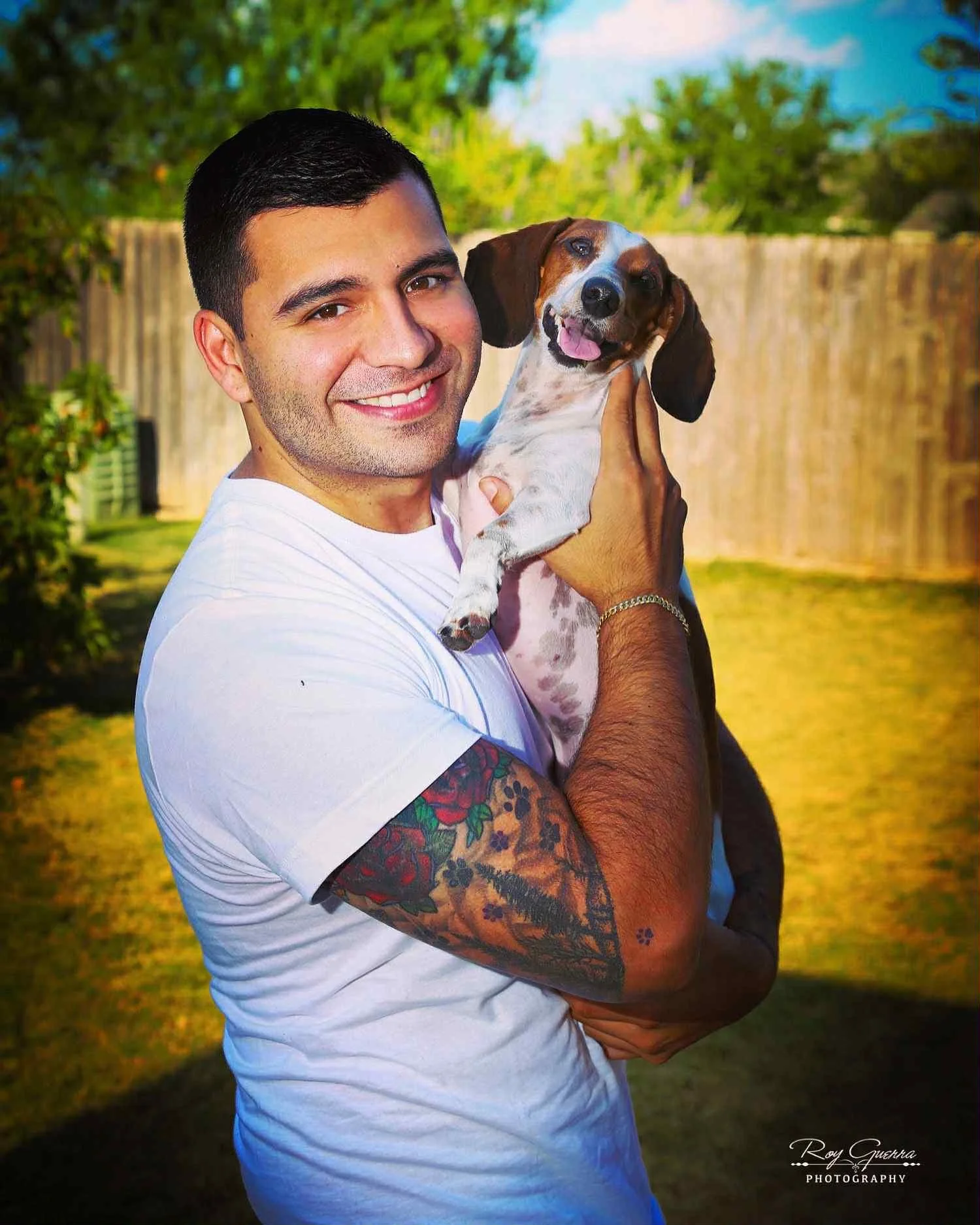 A man with short dark hair, tattoos on his right arm, wearing a white T-shirt, smiling and holding a happy small brown and white dog with long ears outdoors in a backyard with a wooden fence, trees, and a blue sky.