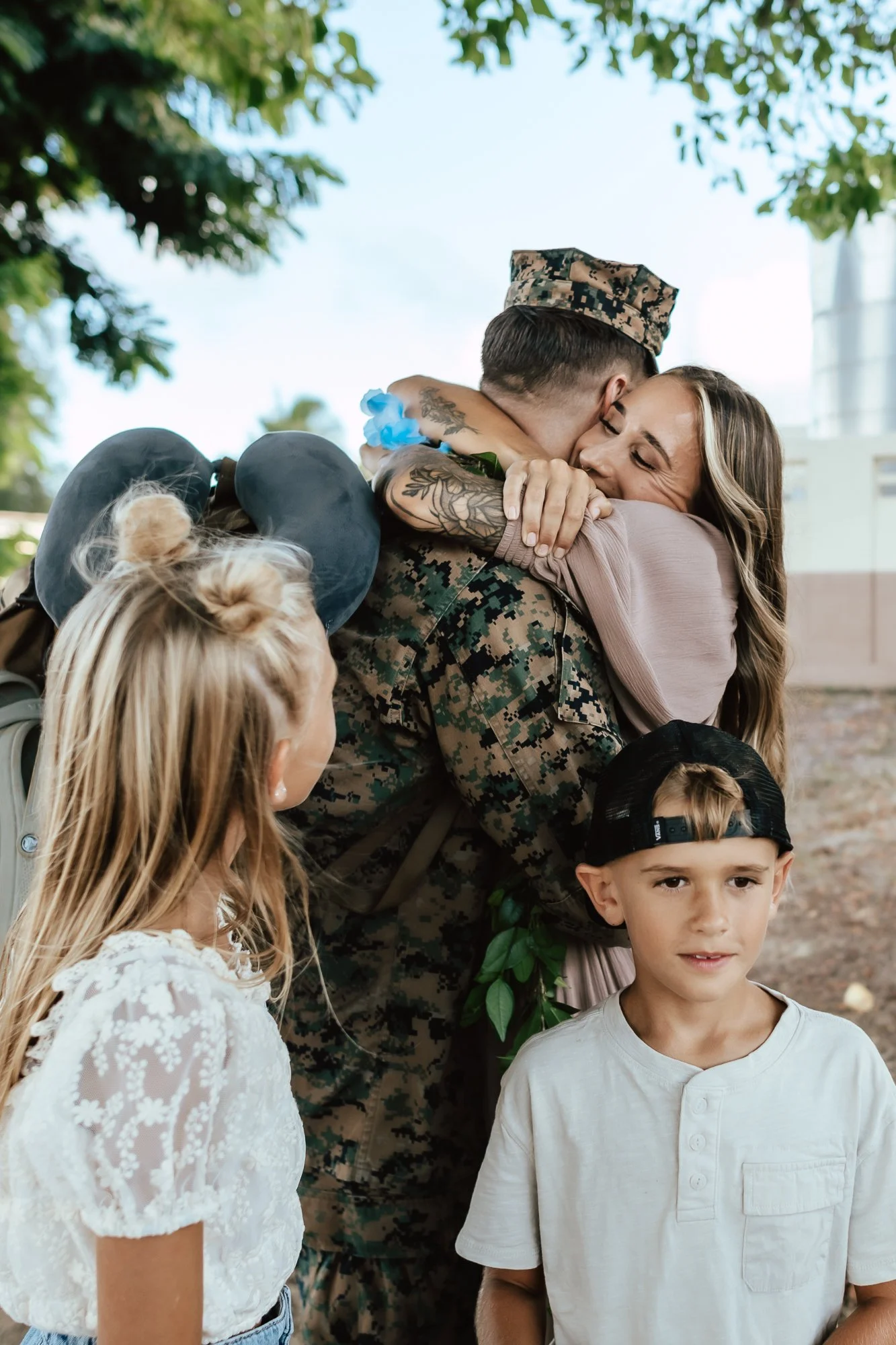 A soldier in uniform hugging and greeting a woman with a big smile, while two children look on. The scene is outdoors with trees and a building in the background. San Antonio Military Homecoming Photographer