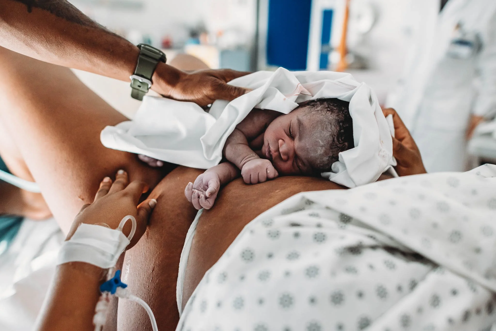 Newborn baby lying on mother’s chest in hospital, wrapped in white cloth, with medical equipment visible in hospital background.