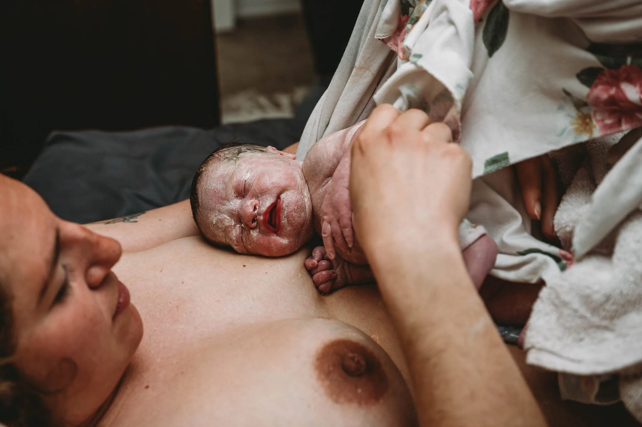 A woman has just given birth. She is lying on her back, holding her newborn baby on her chest. The baby is covered in vernix and appears to be crying or making sounds, with some amniotic fluid on its skin. The woman is looking at the baby lovingly.