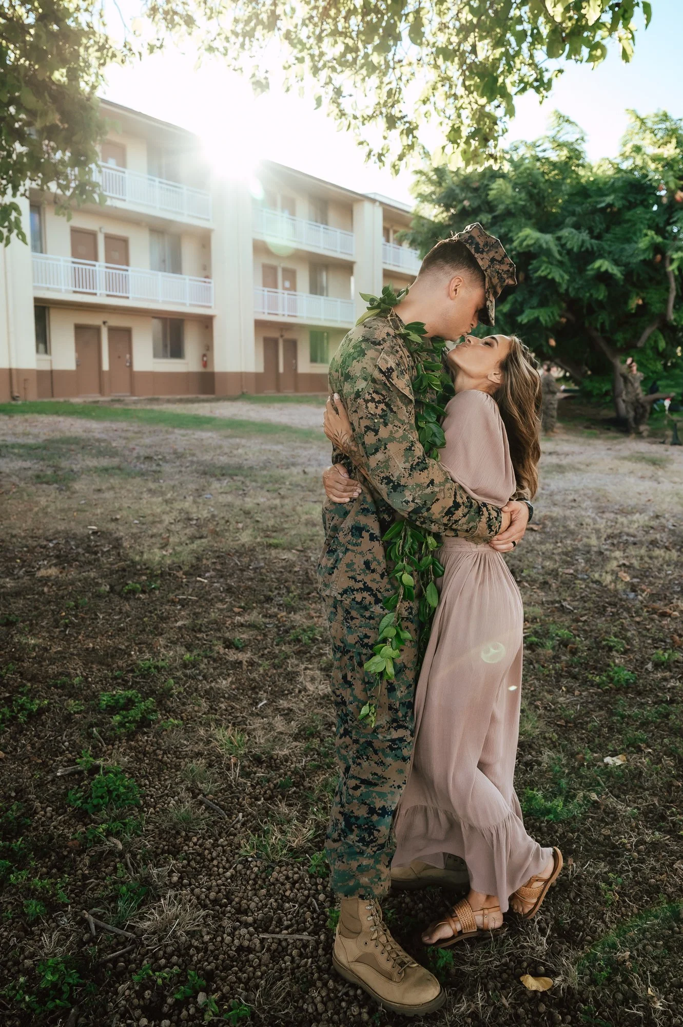 A soldier in camouflage uniform and a woman in a light pink dress are embracing outdoors, with the sun shining behind them and a building with balconies in the background. San Antonio Military Homecoming Photographer