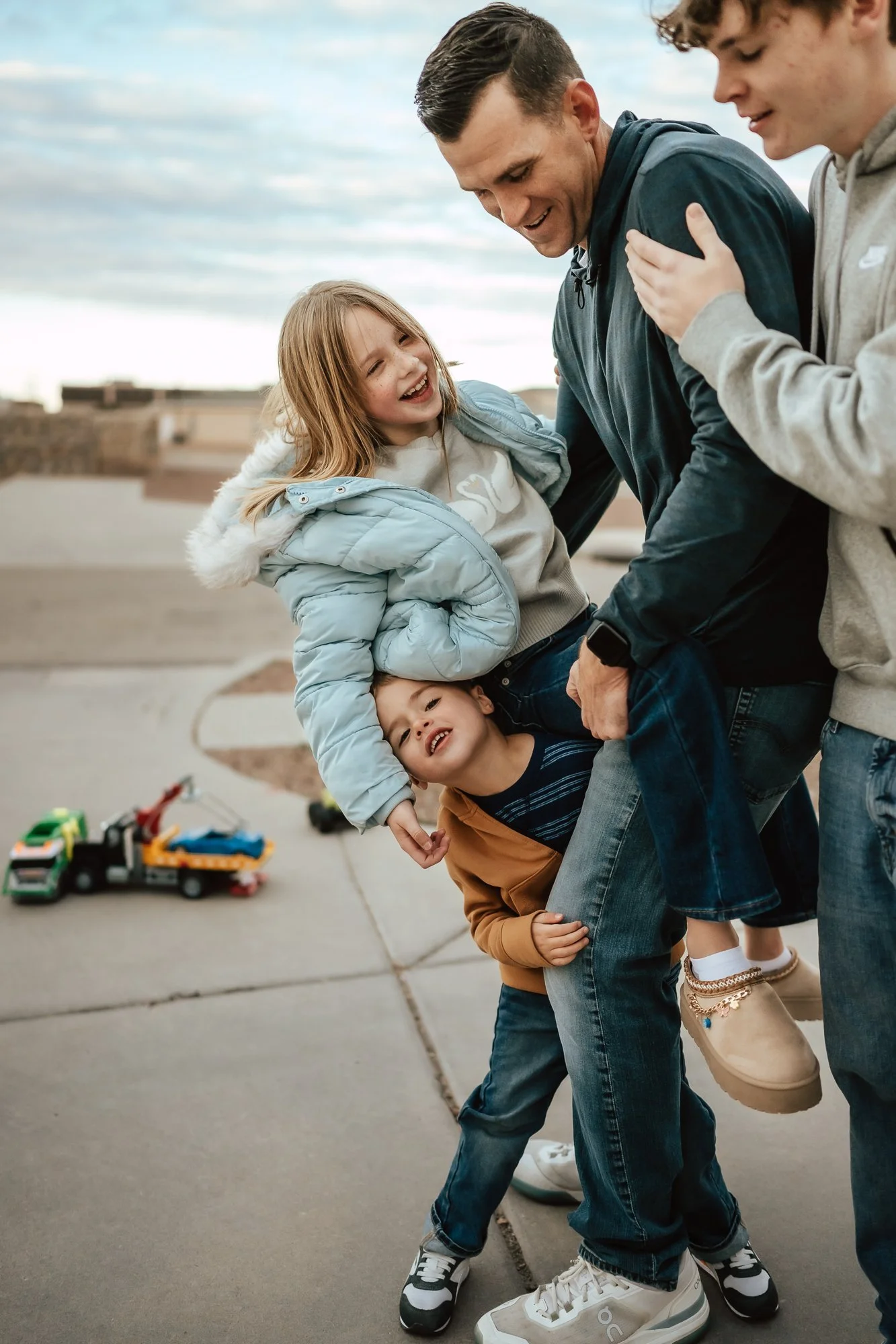 A man playing and being tickled by two children on a sidewalk outdoors, with a girl and a boy smiling and laughing.  San Antonio Family Photographer
