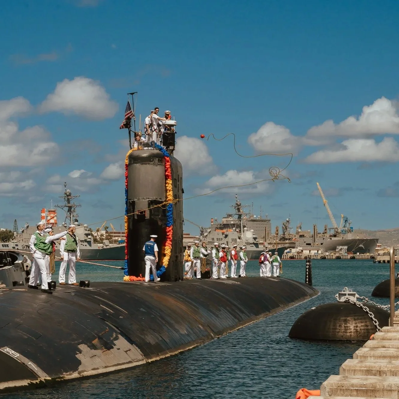A naval crew in white uniforms and colorful vests working on a submarine decorated with flower garlands at a dock, with ships and a crane in the background on a sunny day. San Antonio Military Homecoming Photographer