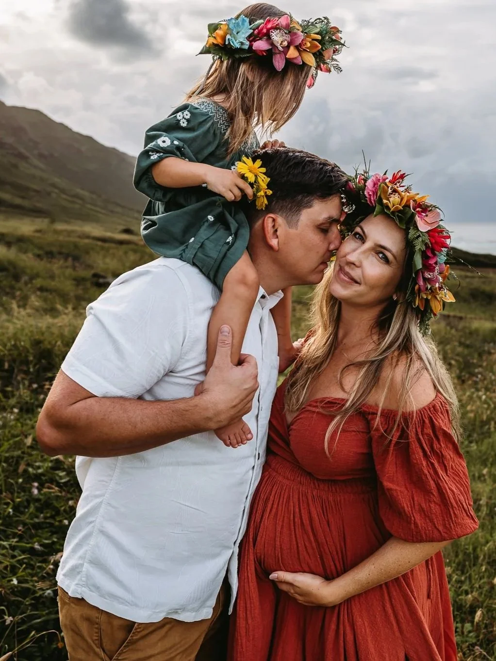A family in a scenic outdoor setting, with a woman in a red dress and flower crown, a man kissing her on the head, a child sitting on his shoulders wearing a green dress and flower crown, near a grassy field and ocean.