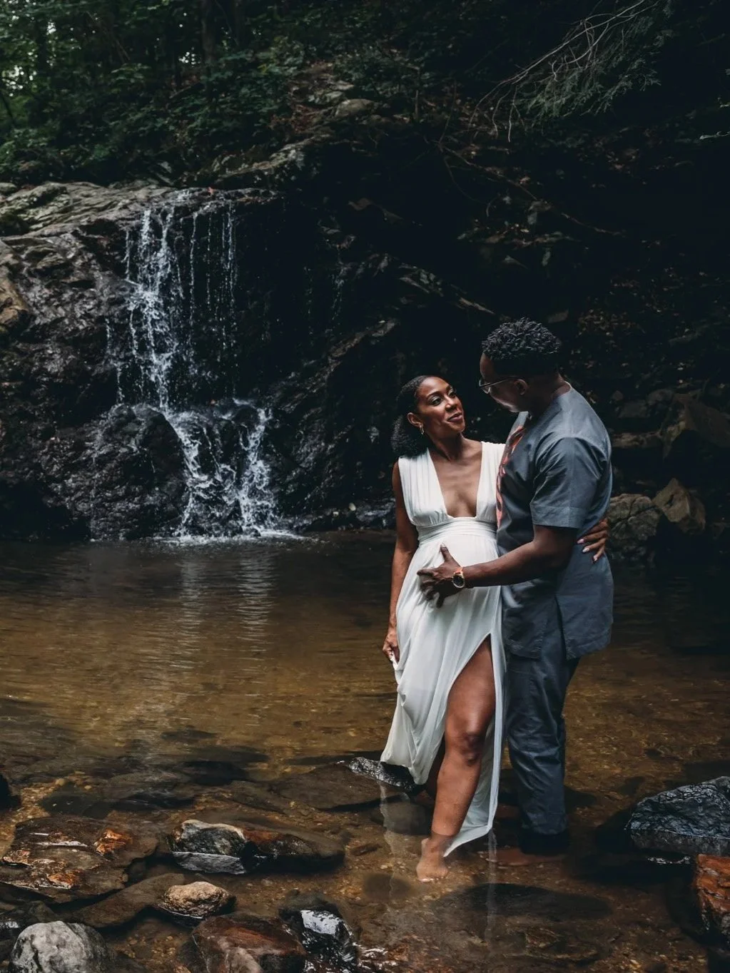 Couple standing in a rocky stream with a waterfall, surrounded by forest.