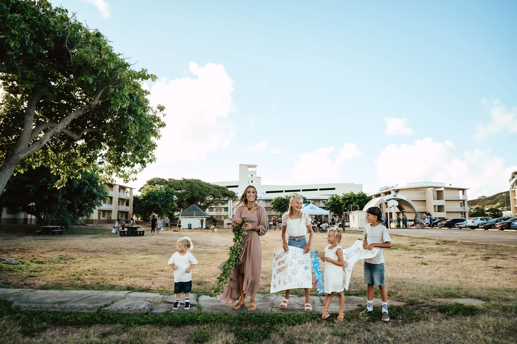 Group of children and two women standing on a grassy field near a school or residential complex, holding welcome signs and banners, during a sunny day. San Antonio Military Homecoming Photographer
