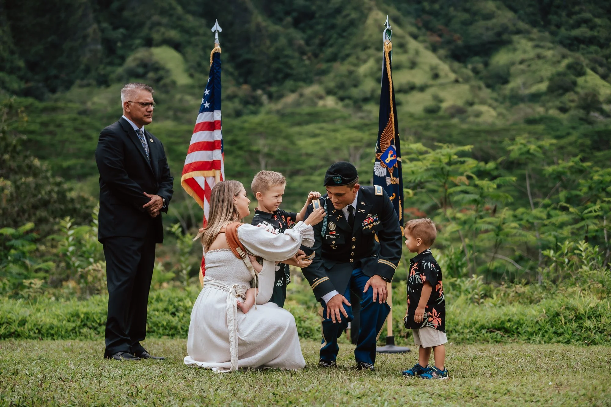 A military promotion ceremony outdoors with lush green hills in the background. A wife pins rank to a man in a military uniform, surrounded by children and two flags, the American flag and a military flag. San Antonio Military Event Photographer