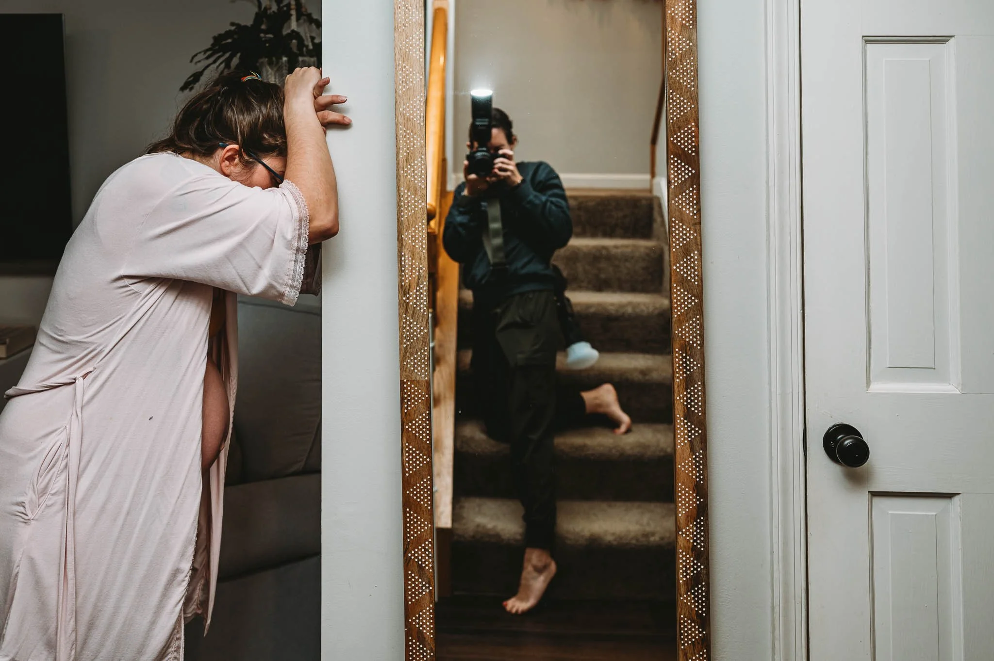 Person in pink pajamas leaning against a wall with eyes closed, looking downwards. Their hand rests on their head. A tall mirror reflects a person with a camera taking a photo, dressed in black, kneeling on carpeted stairs.