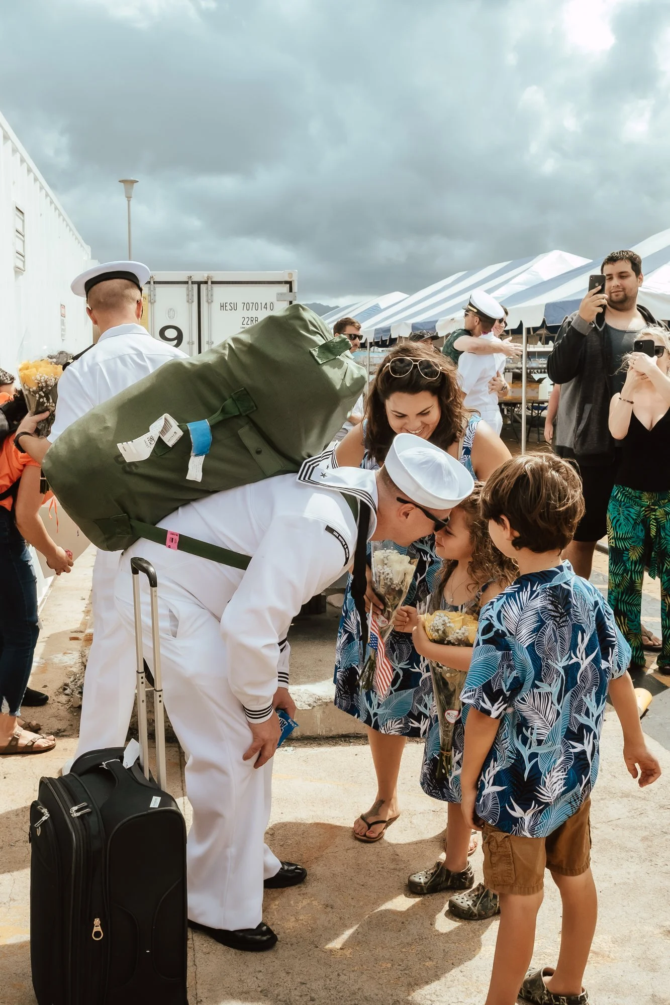 A person dressed in a United States Navy uniform is greeting children holding flowers at an outdoor event, with family members and other sailors nearby under a cloudy sky. San Antonio Military Homecoming Photographer