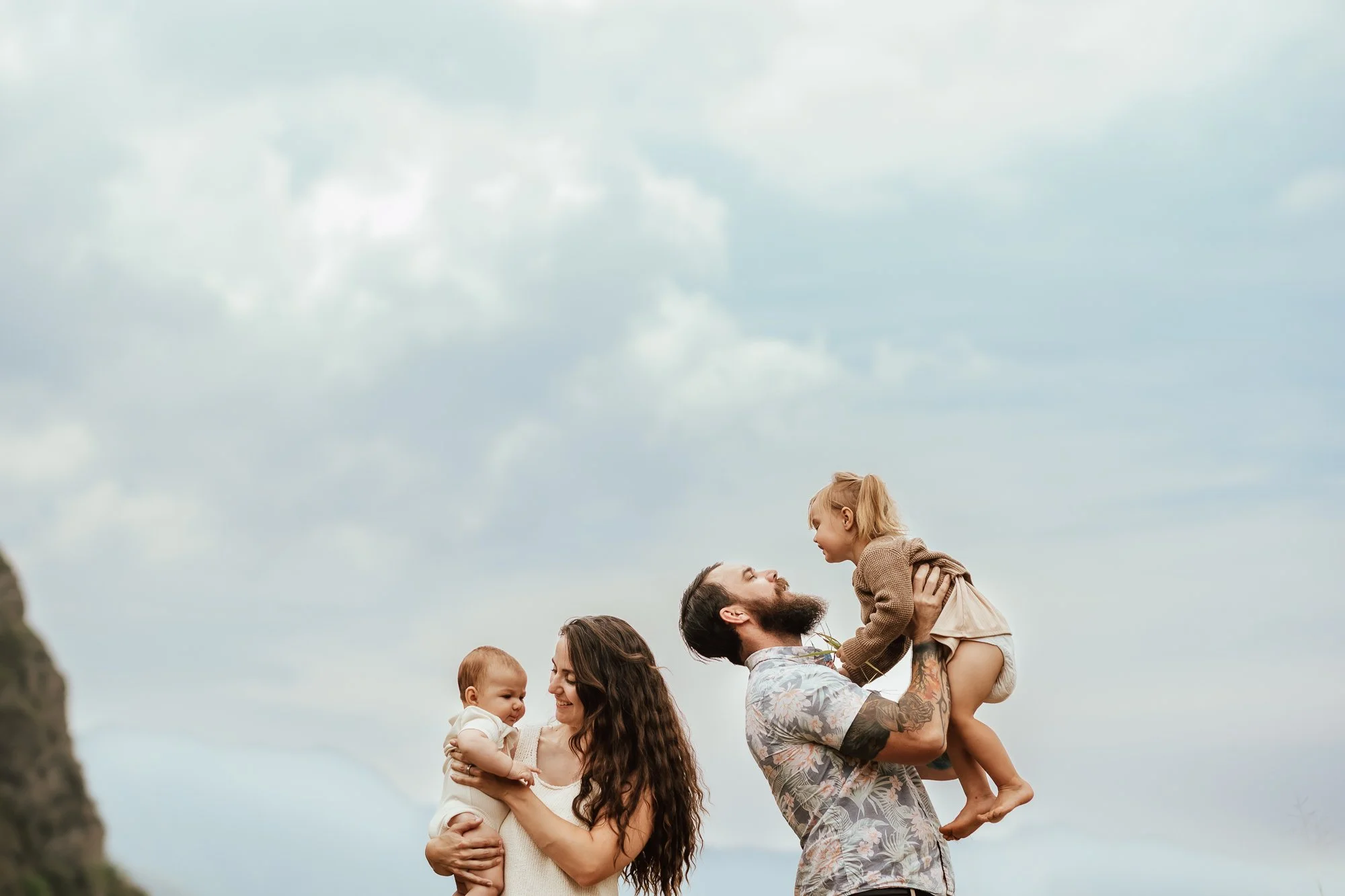 A family of four outdoors on a cloudy day, with a woman holding a baby, a man lifting a young girl, and both parents smiling. San Antonio Family Photographer