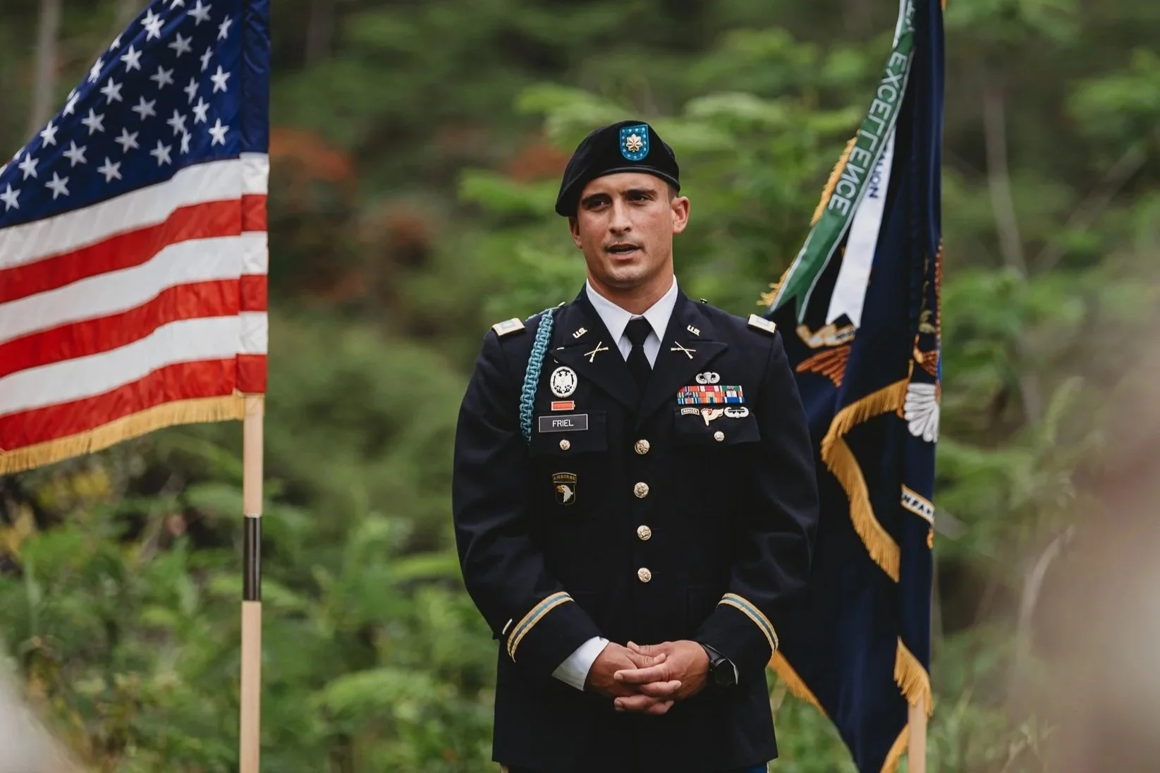 A man in a United States Army uniform standing outdoors with flags on either side, surrounded by green trees. San Antonio Military Event Photographer