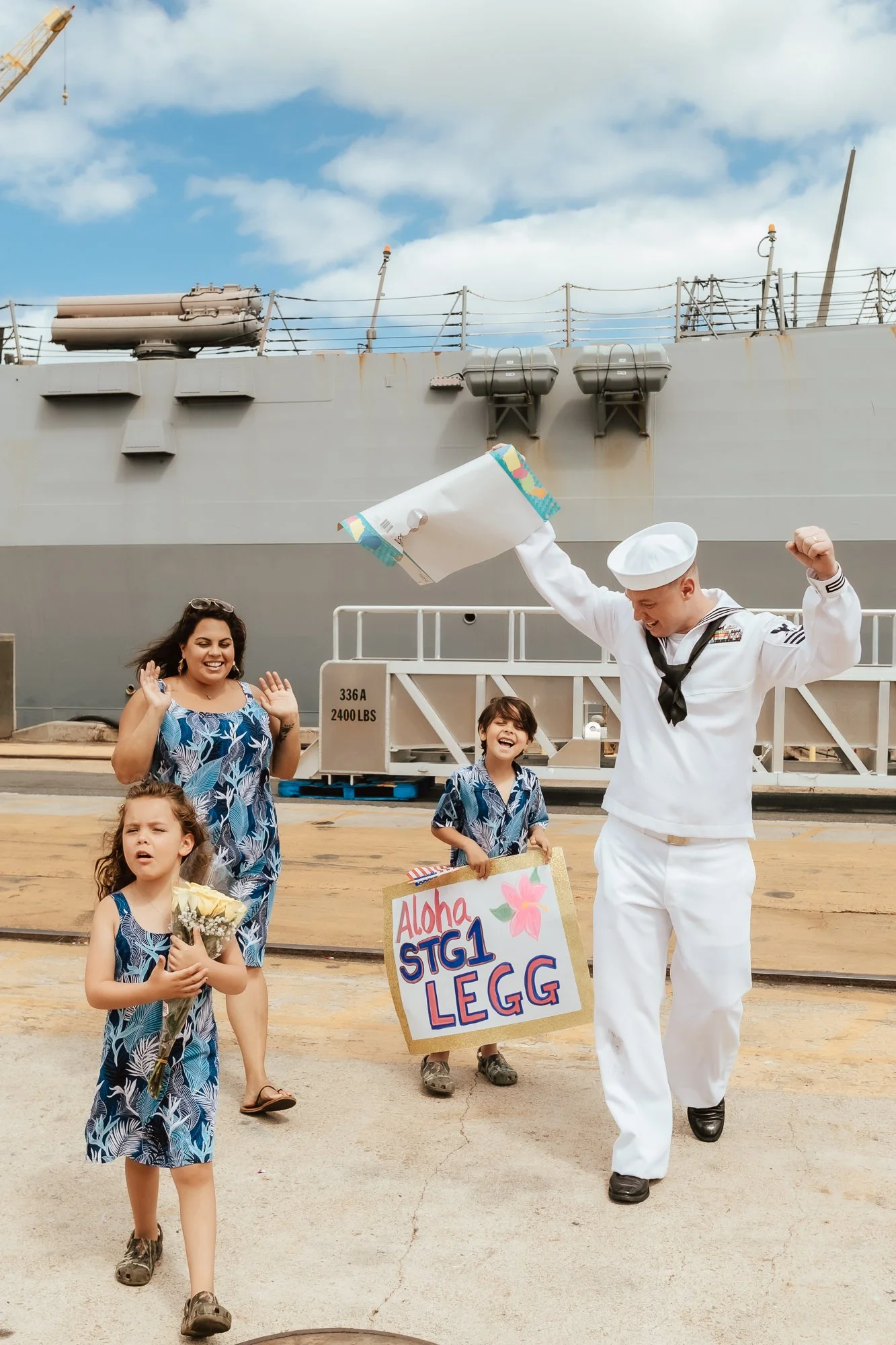 A sailor in uniform celebrating with a family on a ship's dock, holding a homecoming sign up in victory. San Antonio Military Homecoming Photographer