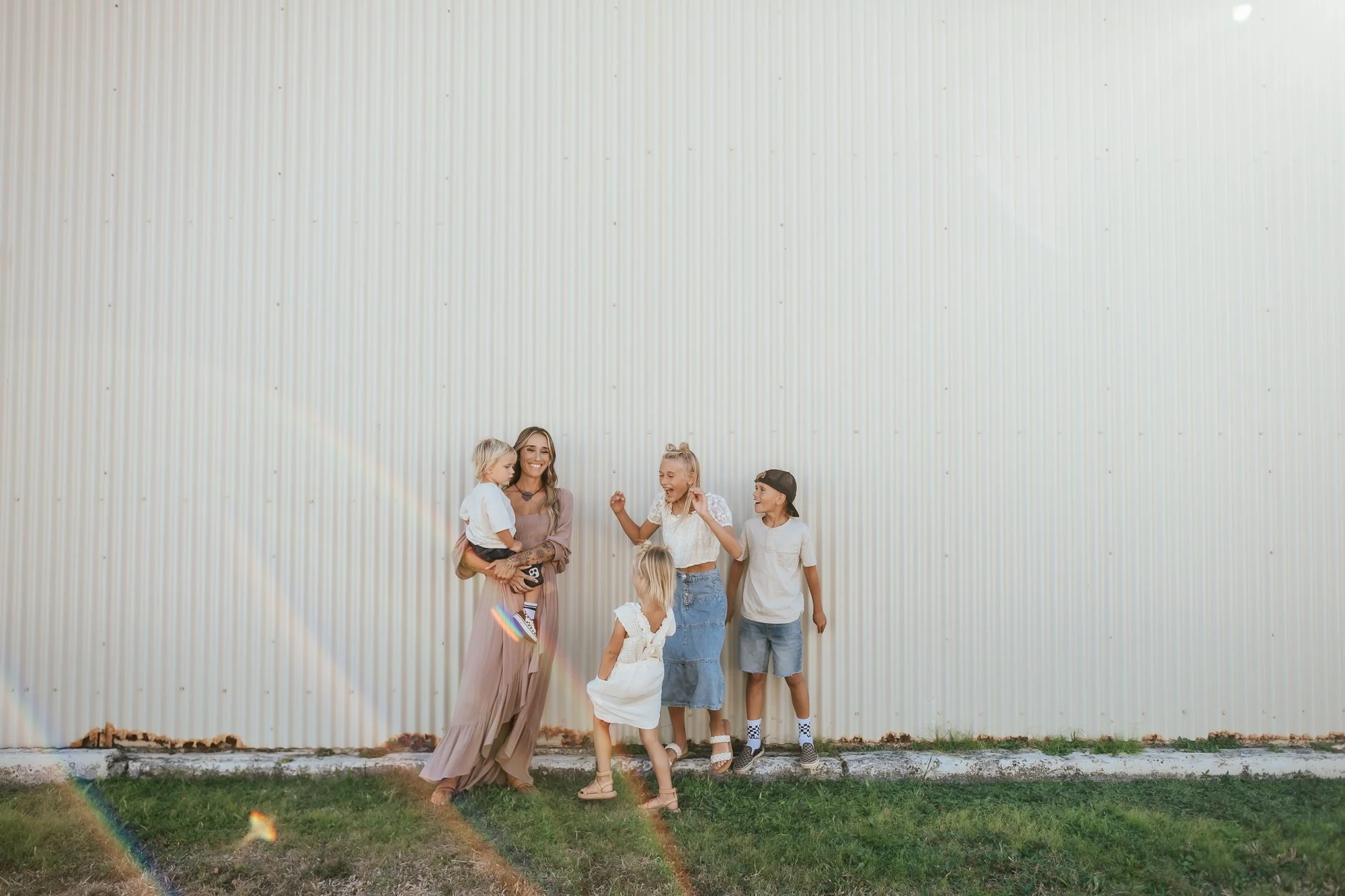 A woman and four children smiling and playing outside near a corrugated metal wall with rainbow light reflections. San Antonio Military Homecoming Photographer