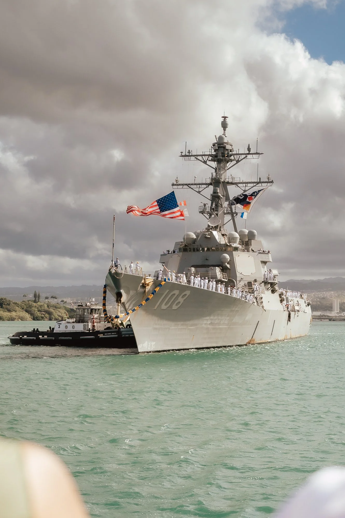 U.S. Navy battleship with flags, crew on deck, sailing in calm water under cloudy sky. San Antonio Military Homecoming Photographer