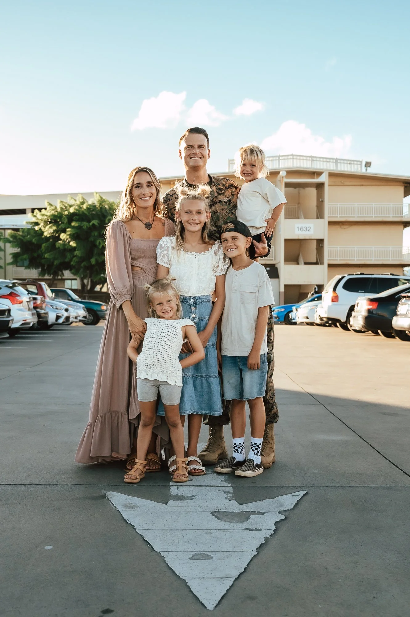 A family of seven, including three kids and four adults, standing together in a parking lot with cars behind them. The group is smiling, and one member in military uniform is holding a young child. San Antonio Military Homecoming Photographer