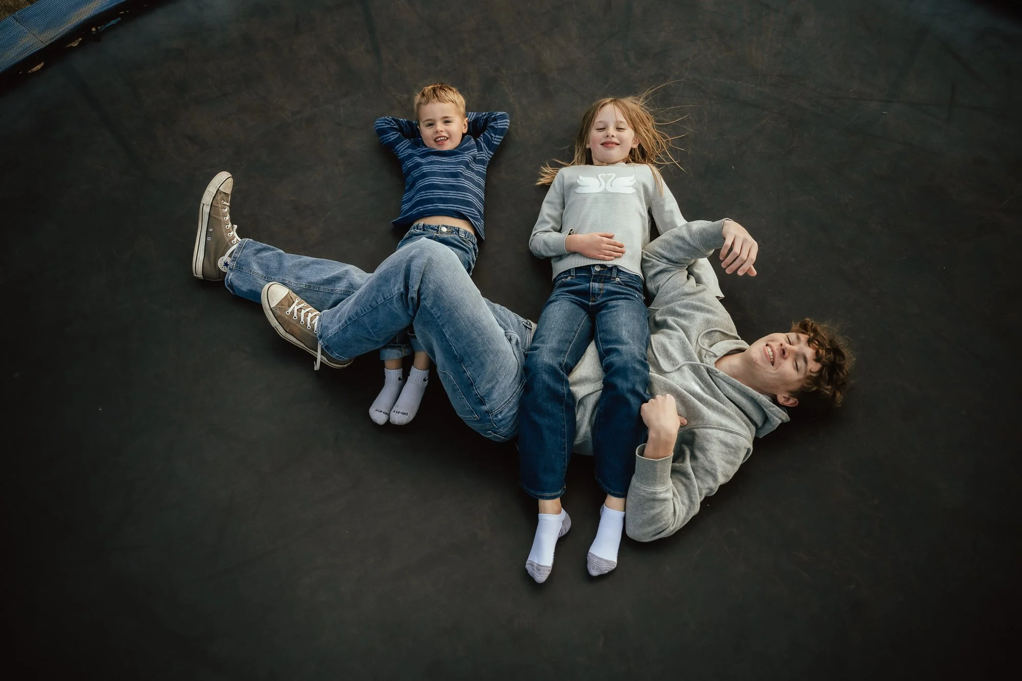 A young man and two children lying on a black mat, smiling and relaxed, with the children positioned on the young man's stomach and chest. San Antonio Family, Newborn and Birth Photographer