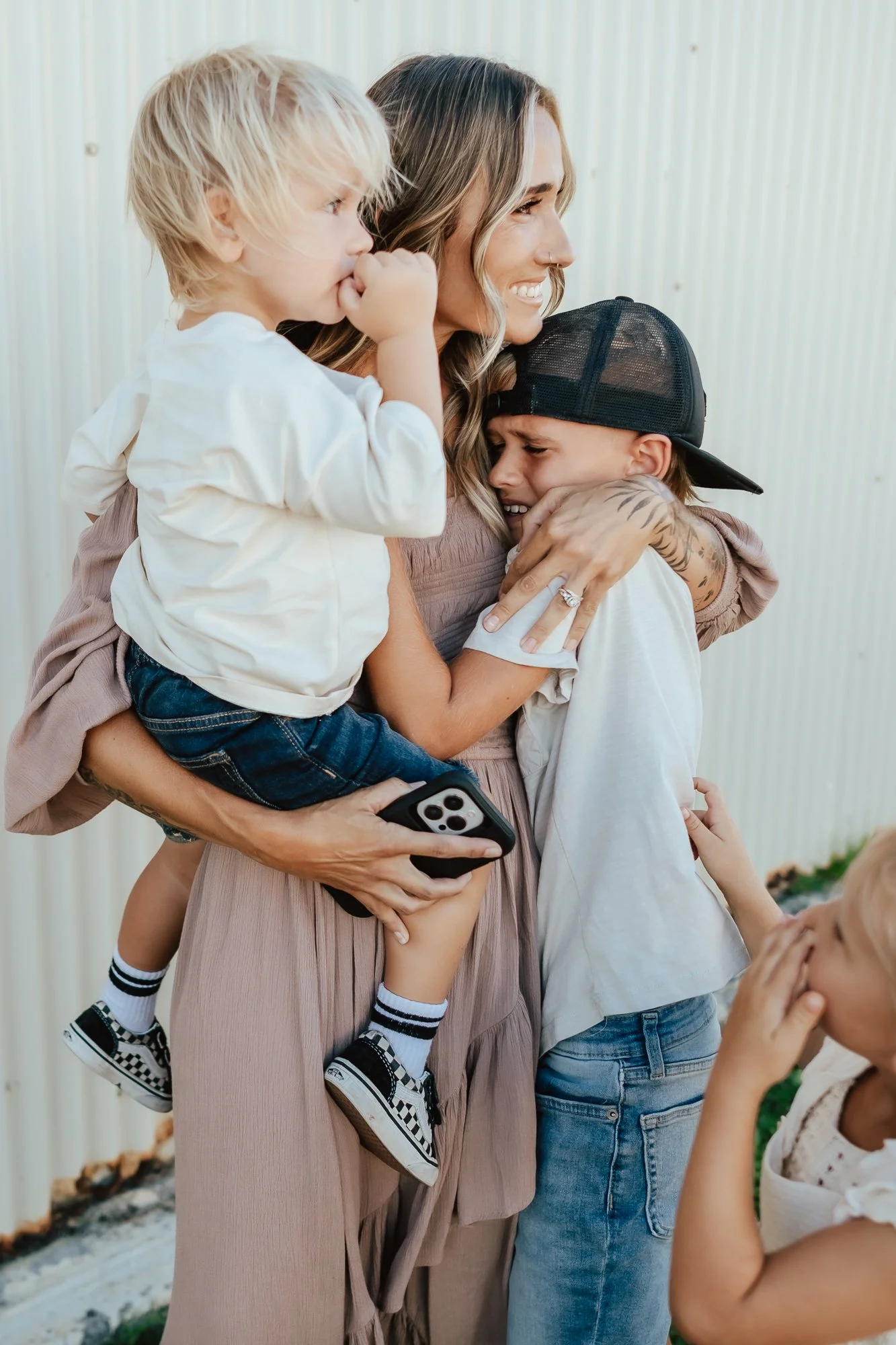A woman is hugging two young boys, one on each side. She is smiling, and the boys are crying as they hug her. The woman is holding a smartphone in her hand. San Antonio Military Homecoming Photographer