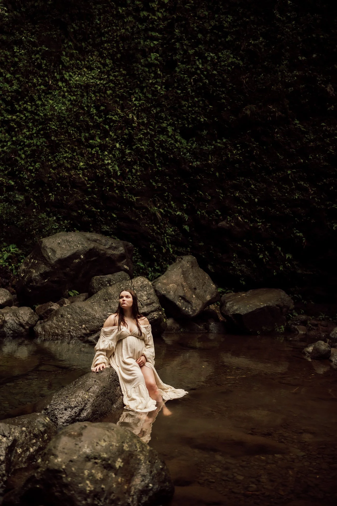 A woman in a beige dress sitting on a large rock in a shallow creek, surrounded by other rocks and a dark green, leafy forest backdrop. Maternity raw in nature photography. 