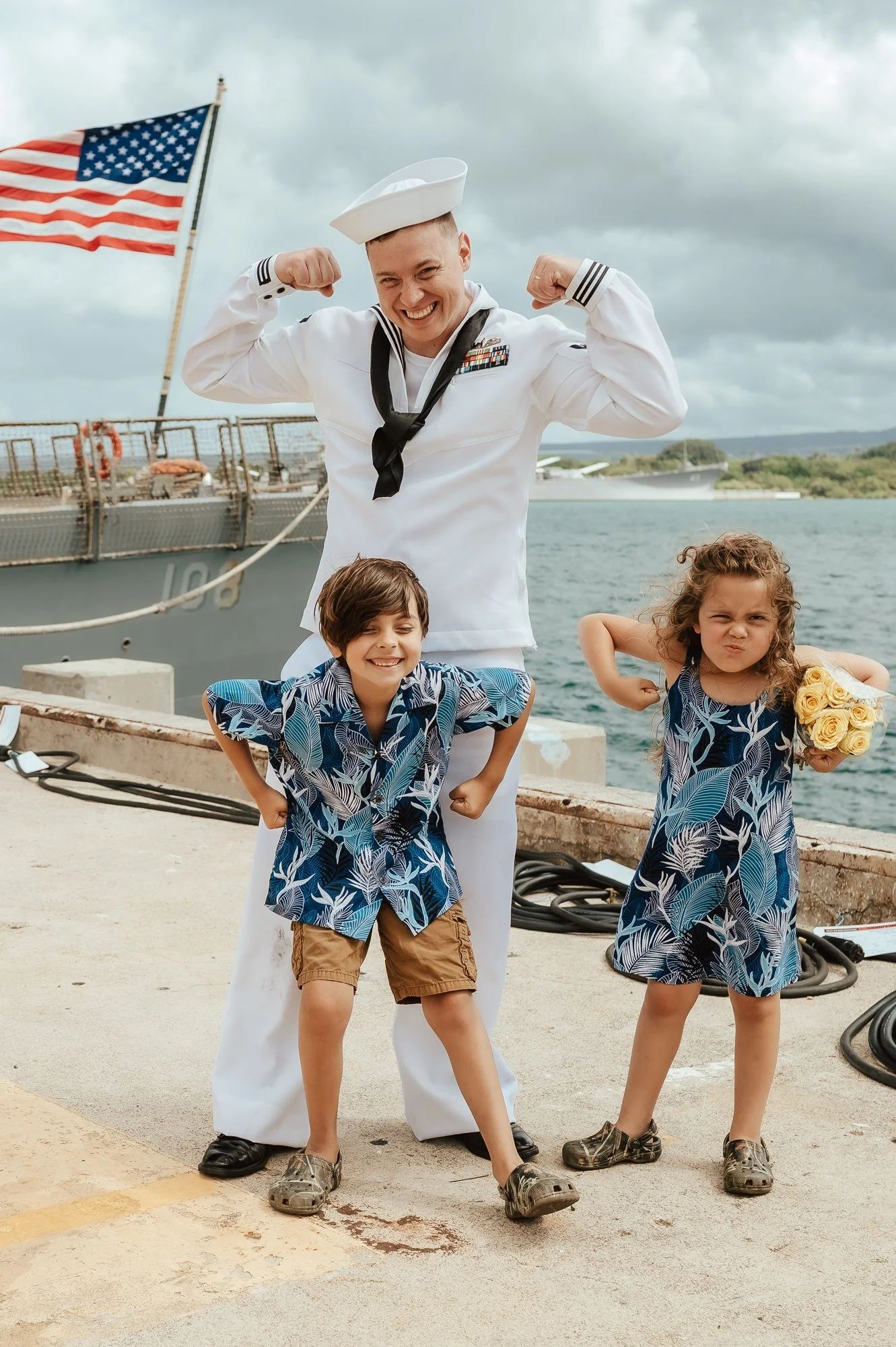 A smiling sailor in a white uniform flexes his arm muscles with two children in matching blue patterned shirts, one boy and one girl, showing strong and playful poses on a dock  with a navy ship behind. San Antonio Military Homecoming Photographer