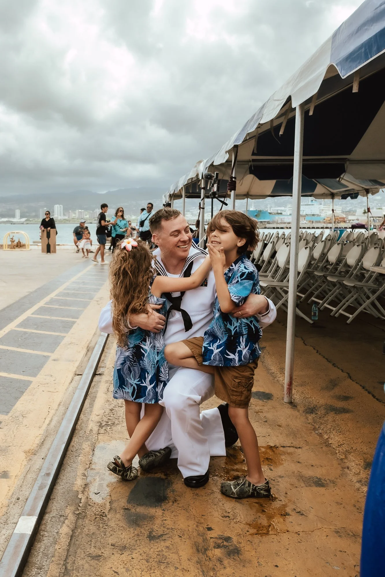 A sailor in uniform happily interacts with two children in matching blue Hawaiian shirts outdoors near a waterfront, with other people and chairs in the background under cloudy skies. San Antonio Military Homecoming Photographer